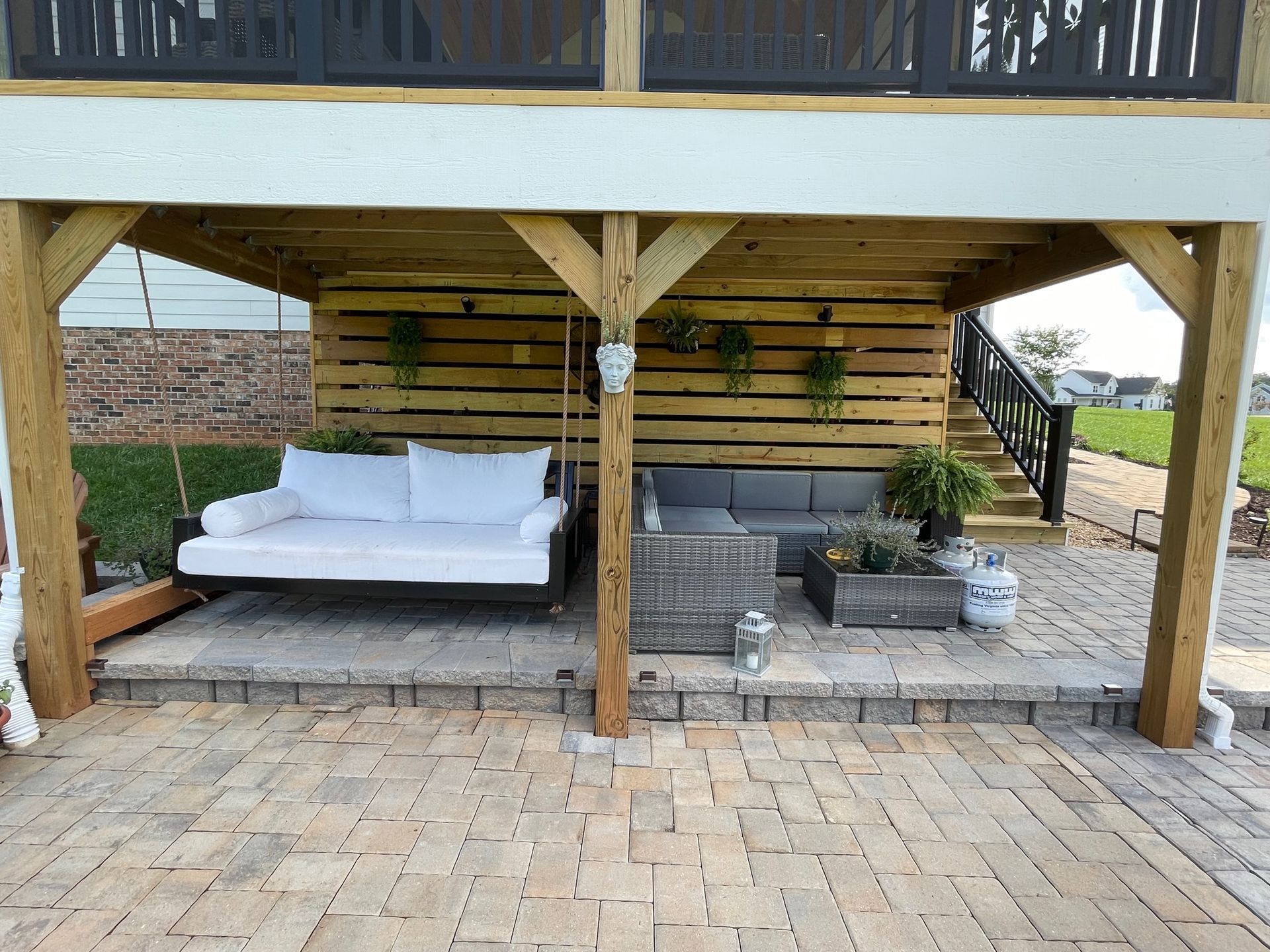 Covered outdoor patio with a white sofa, wooden posts, and potted plants on a brick-paved area