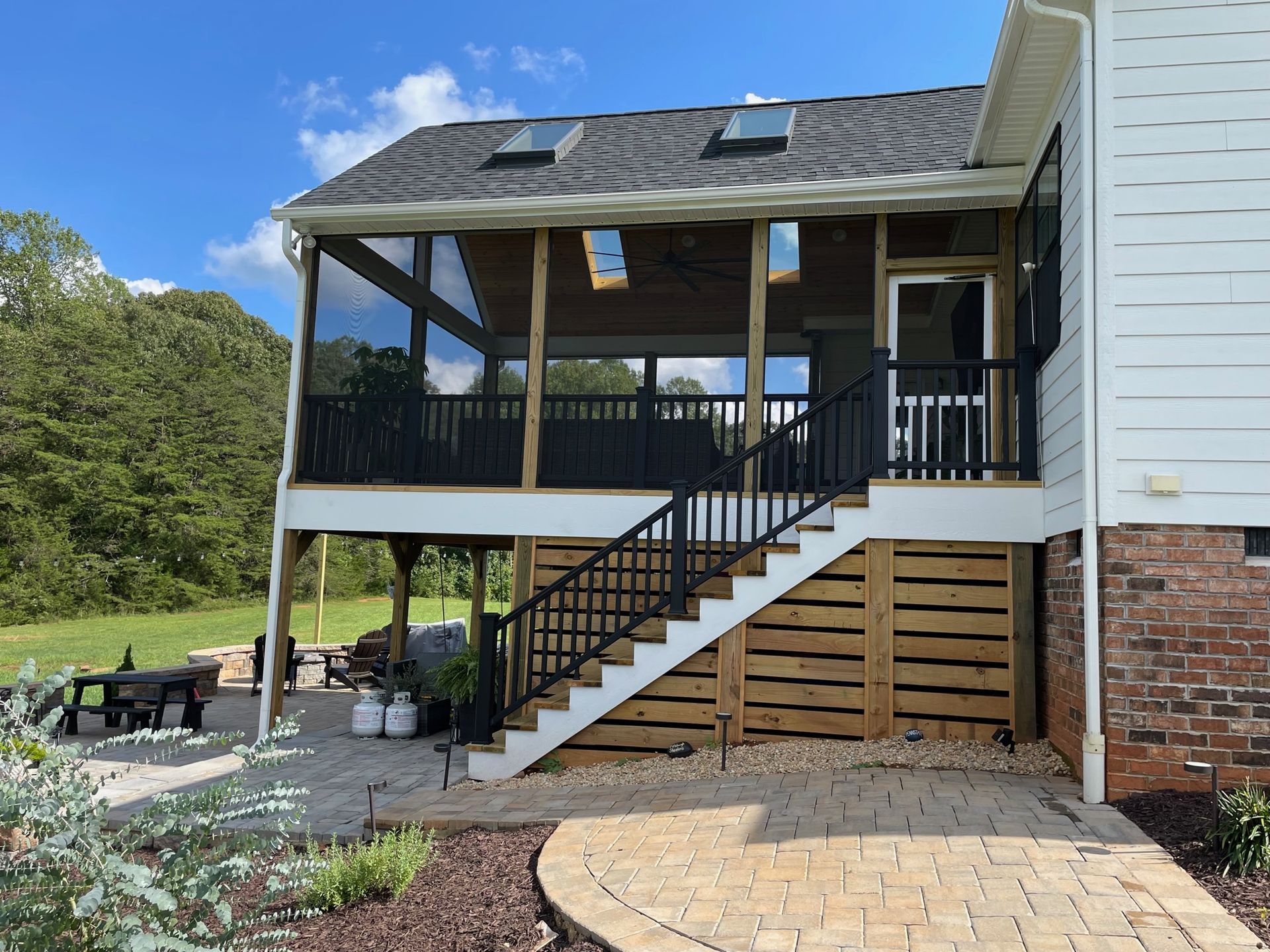 Covered backyard porch with stairs, black railing, brick foundation, and outdoor seating beside a white house.