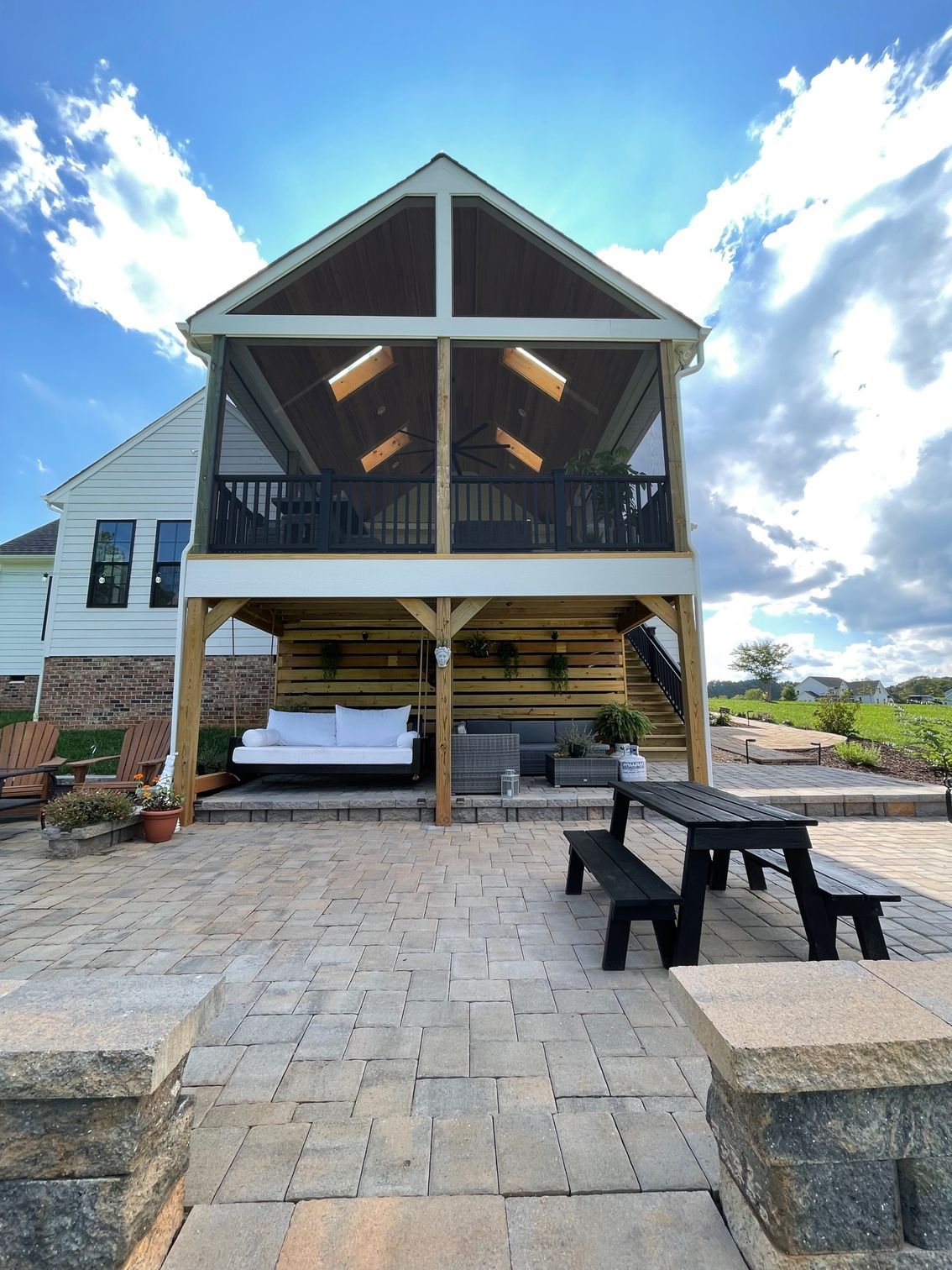 Two-story house with a covered porch, stone patio, and benches under a bright blue sky