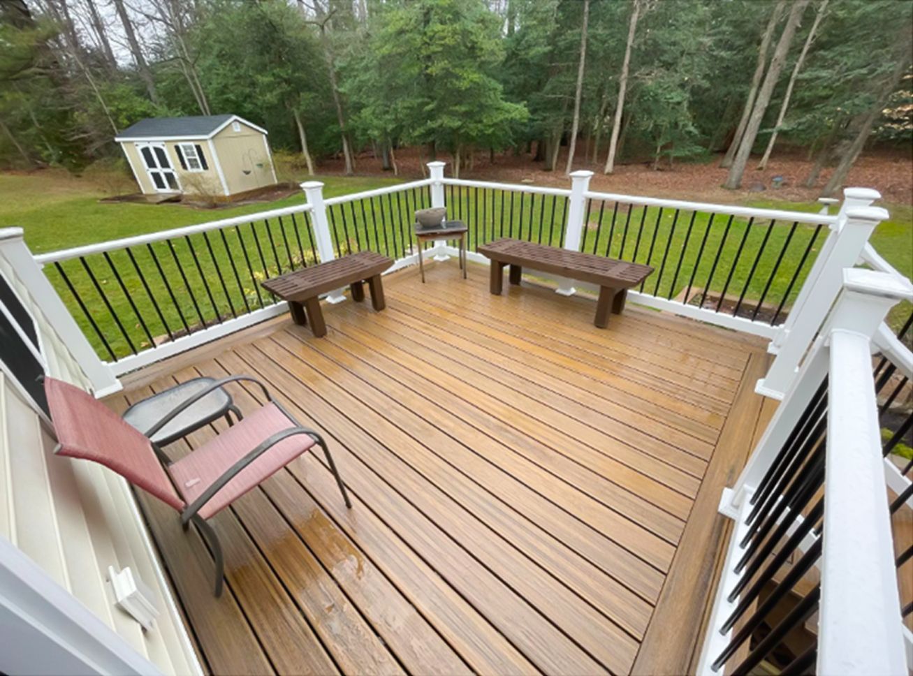 Wooden backyard deck with benches, chair, railing, and grassy yard with shed and trees.