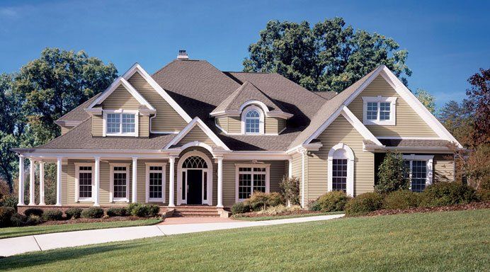 Large beige suburban house with white trim, gabled roofs, and a manicured green lawn under a blue sky