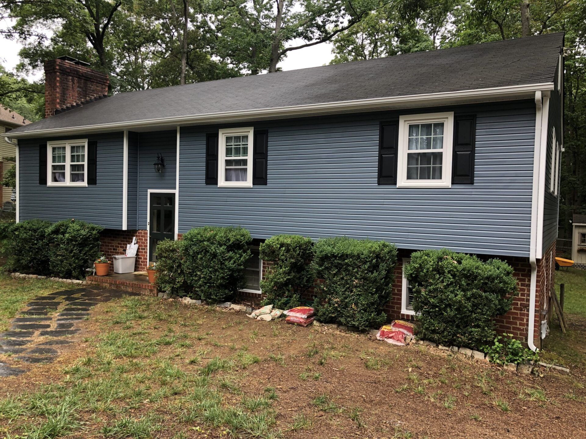 Blue house with white trim, black roof, and shrubs along the front in a wooded yard.