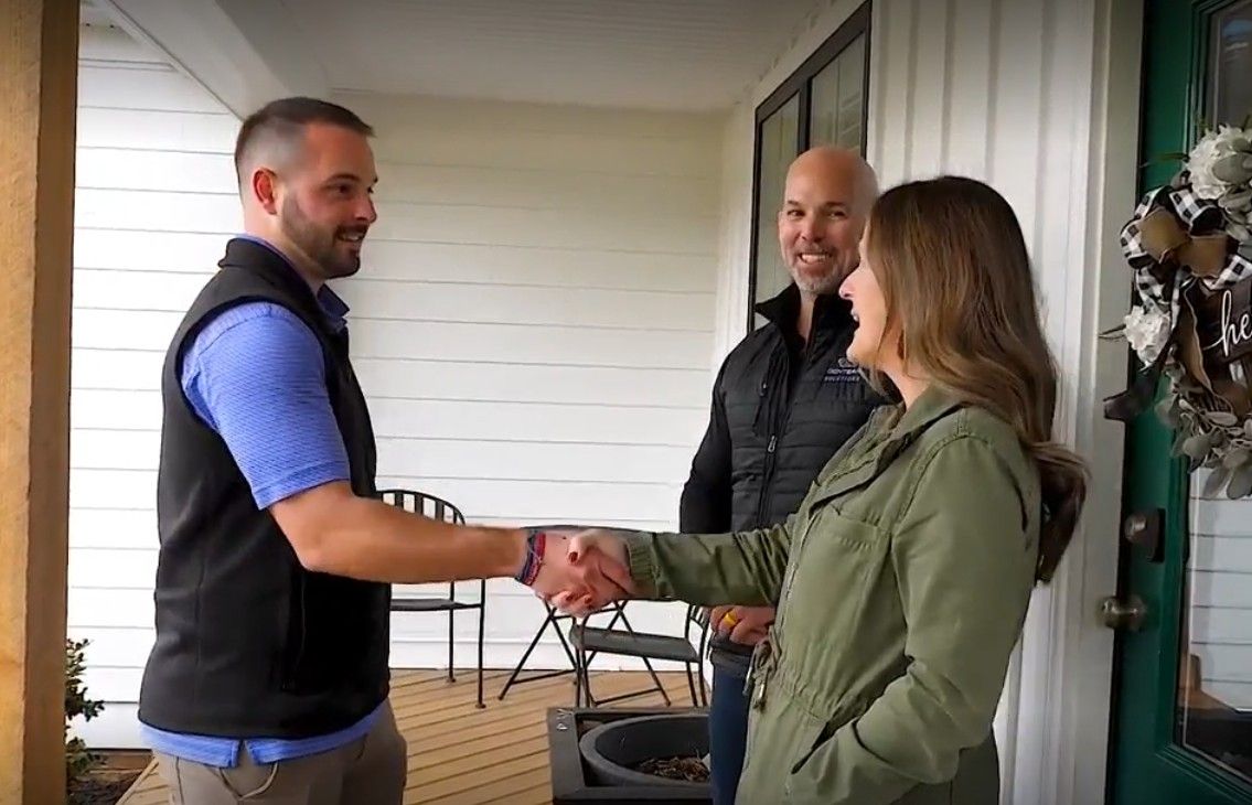 Two people shaking hands on a porch while a third person smiles nearby.