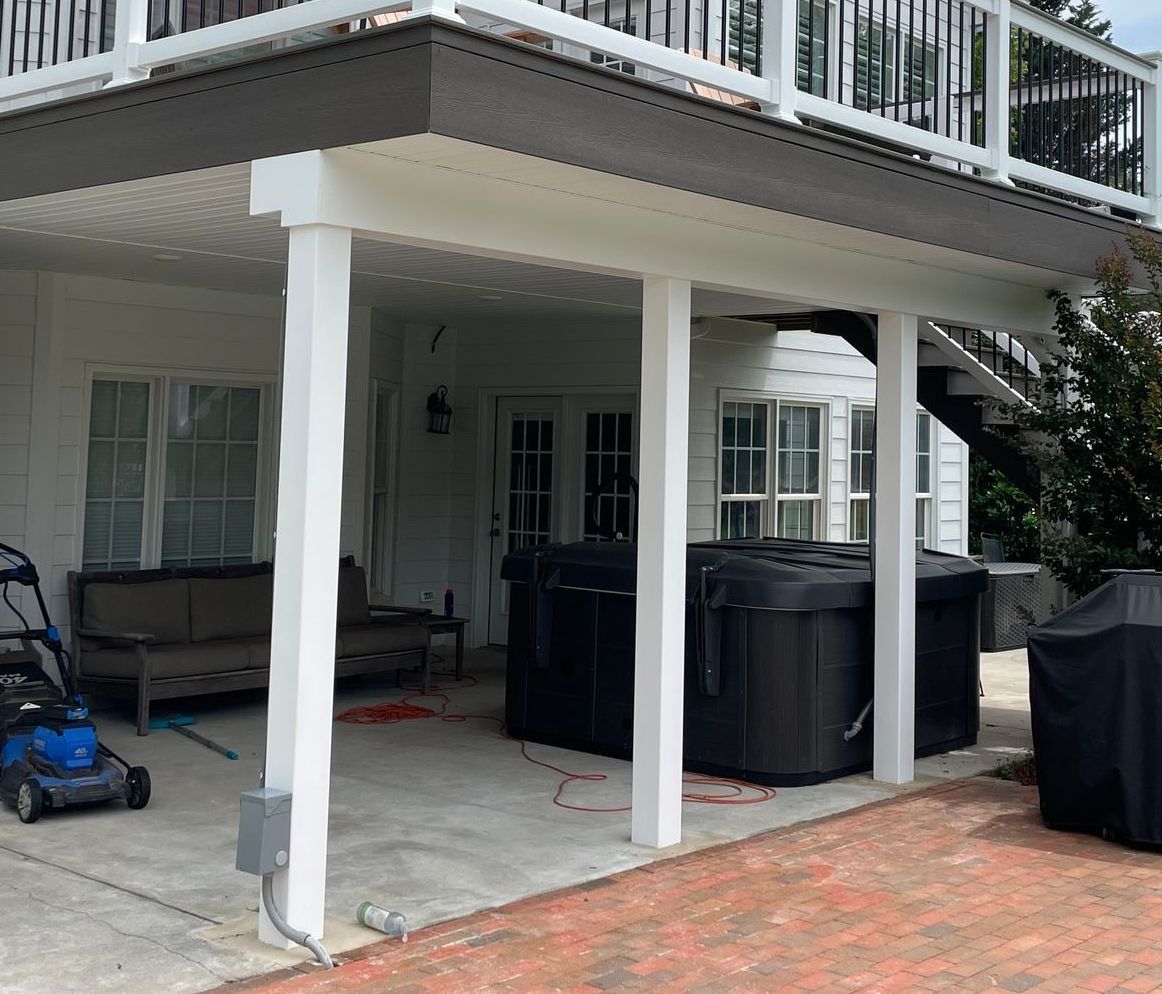 Covered patio under a deck with white columns, black storage bins, and a brick floor.