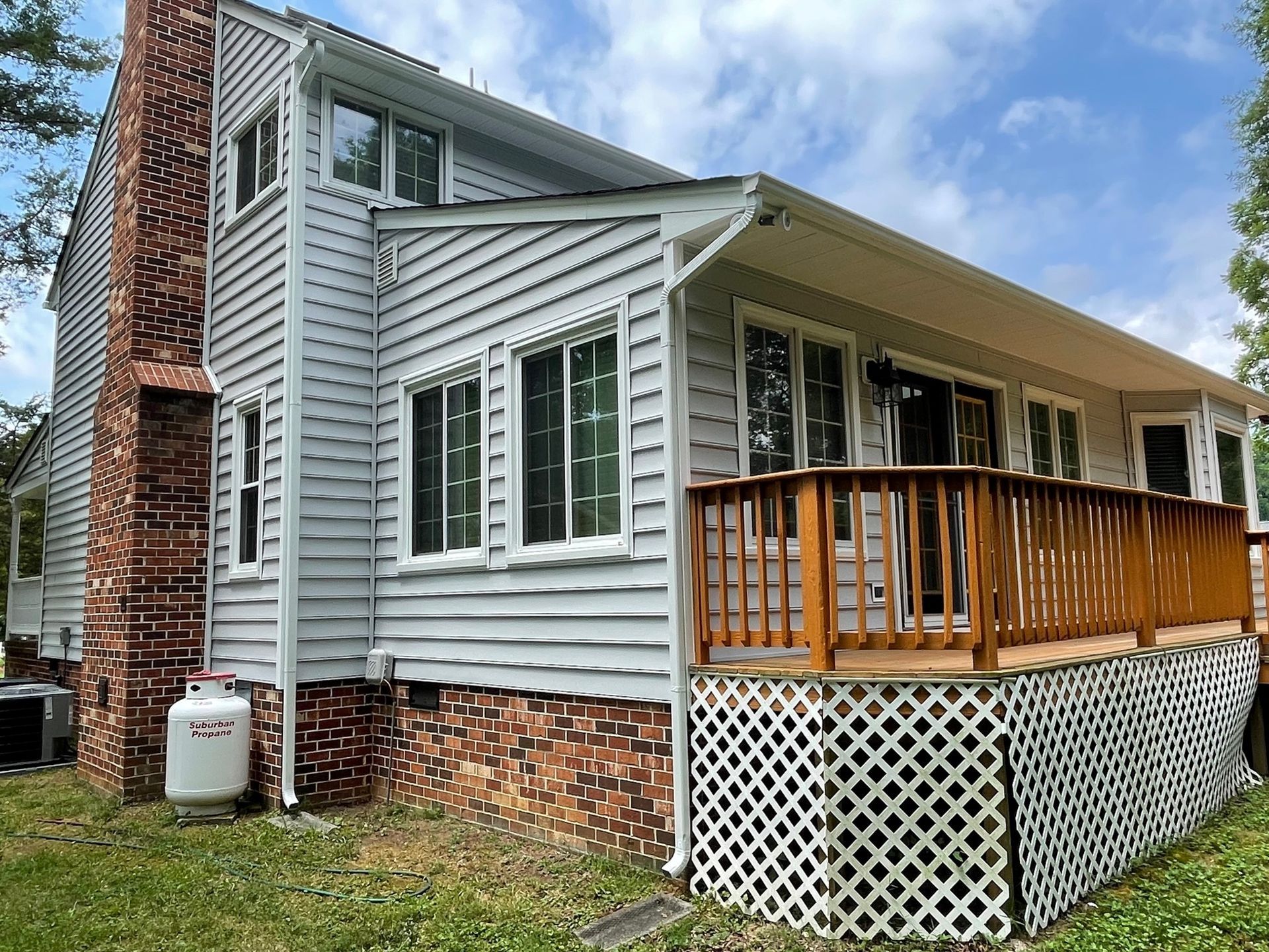 Gray house with brick chimney, wooden porch, and white lattice skirting under a blue sky