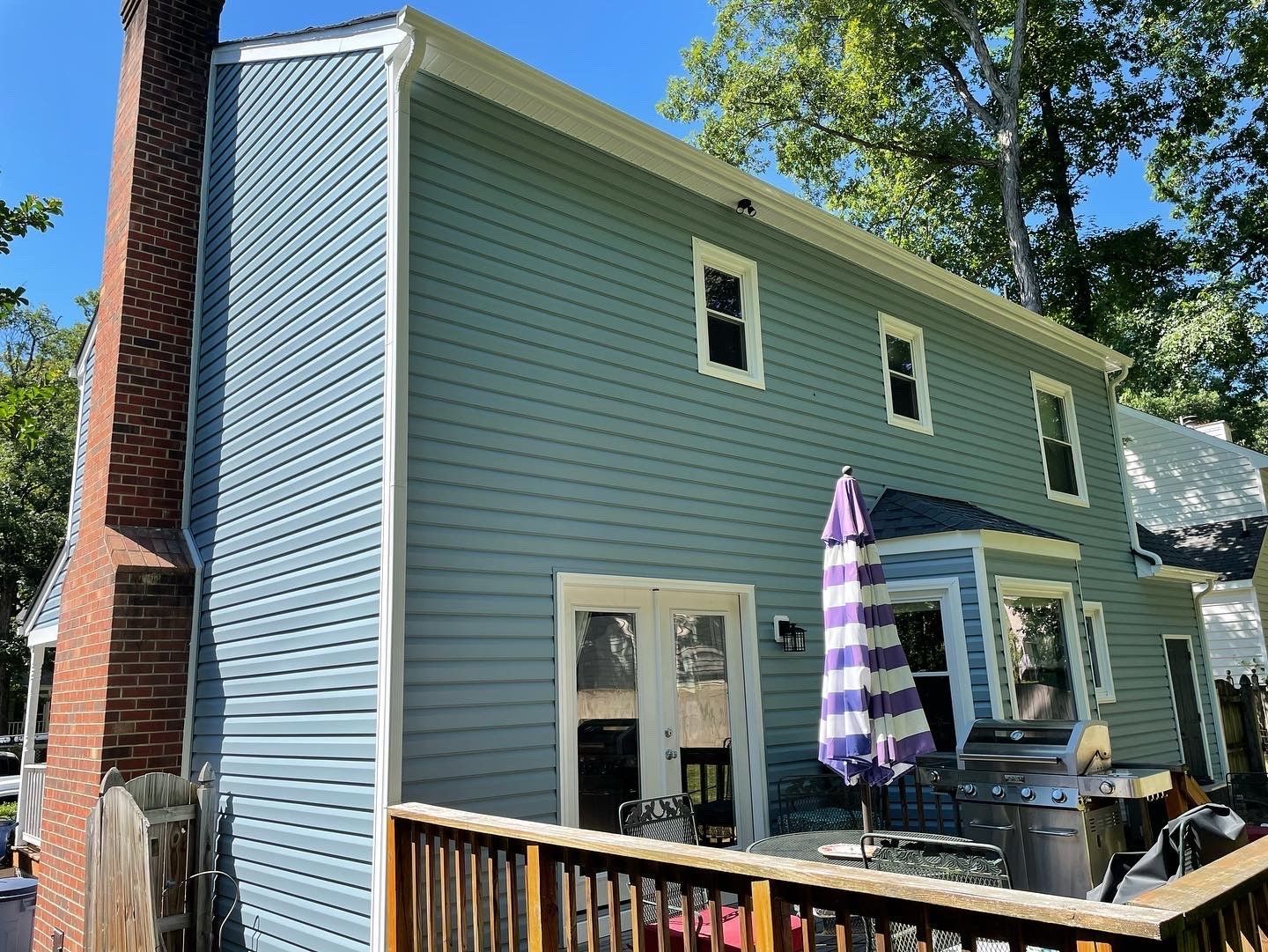 Back patio of a green house with white trim, brick chimney, sliding door, and purple-striped umbrella.