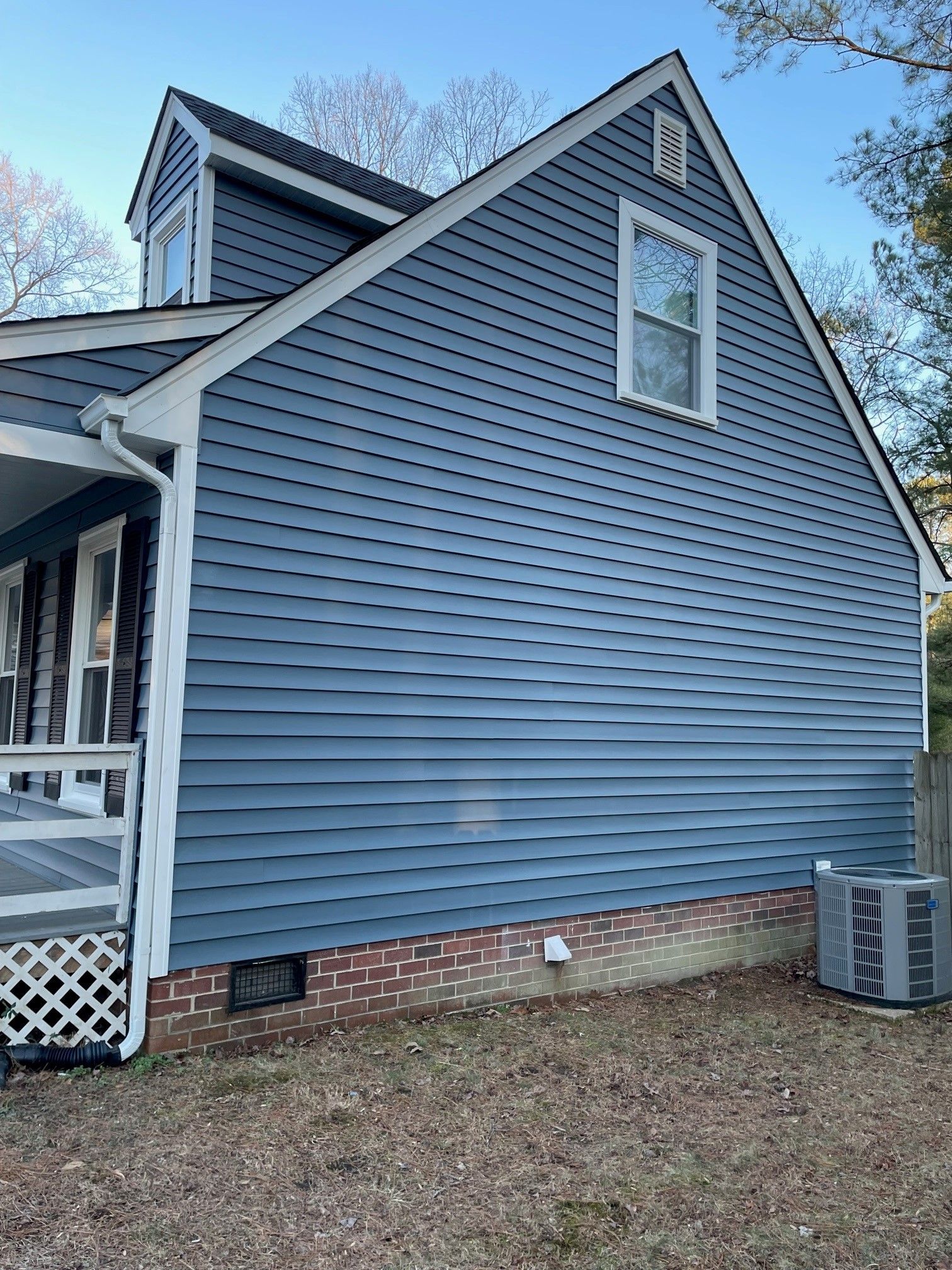 Blue-sided house exterior with brick foundation, porch, and HVAC unit on a grassy lot