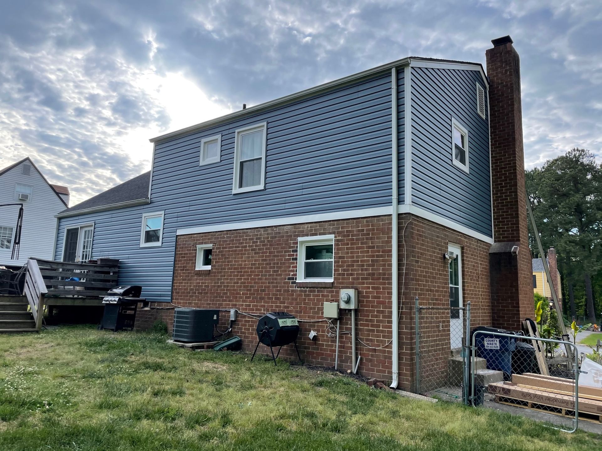 Two-story brick and gray house exterior with chimney, deck, and grassy yard under cloudy sky