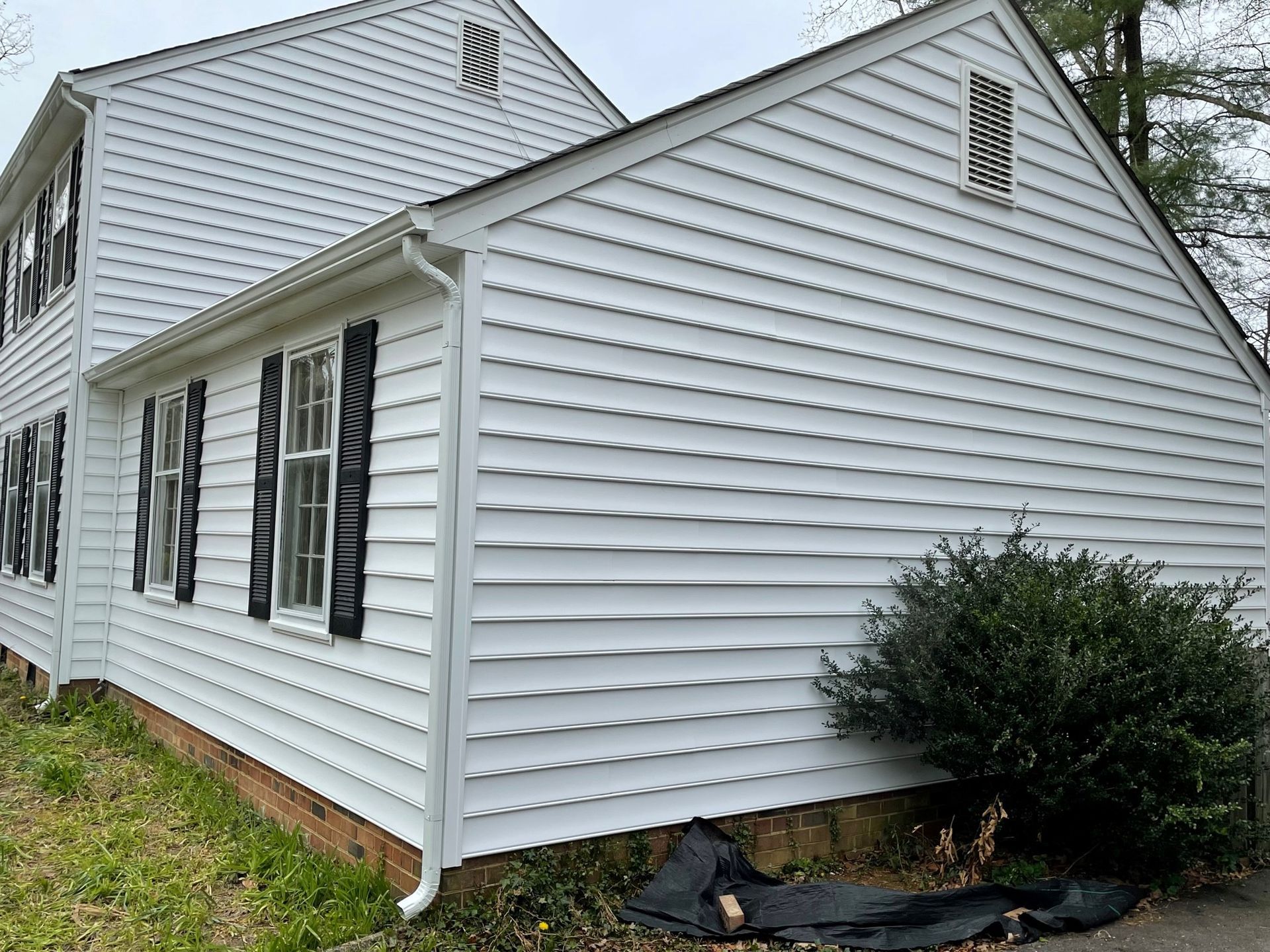 White-sided house exterior with black shutters, a bush by the corner, and some exposed foundation.