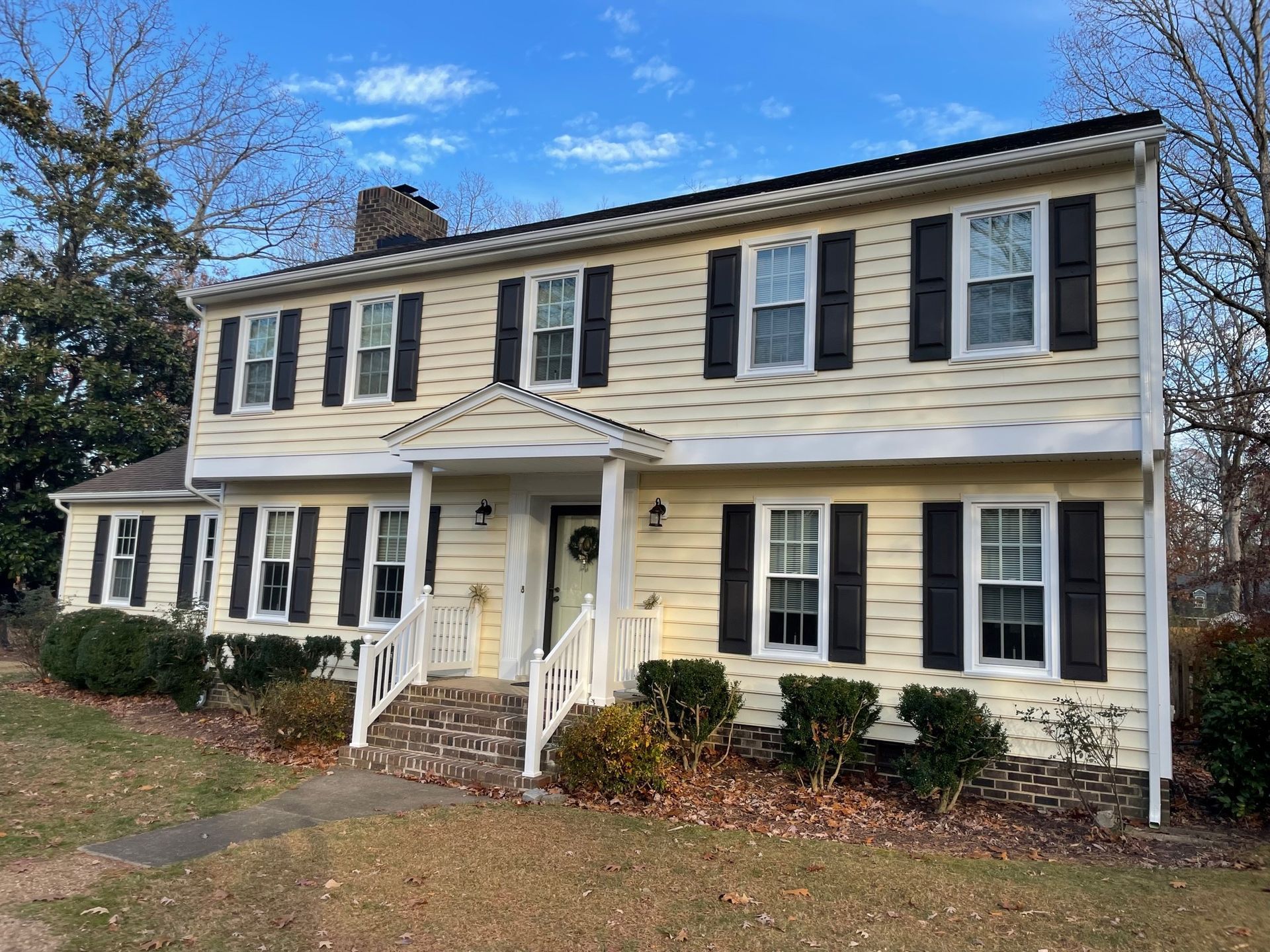 Two-story cream house with black shutters and white porch railings under a blue sky.