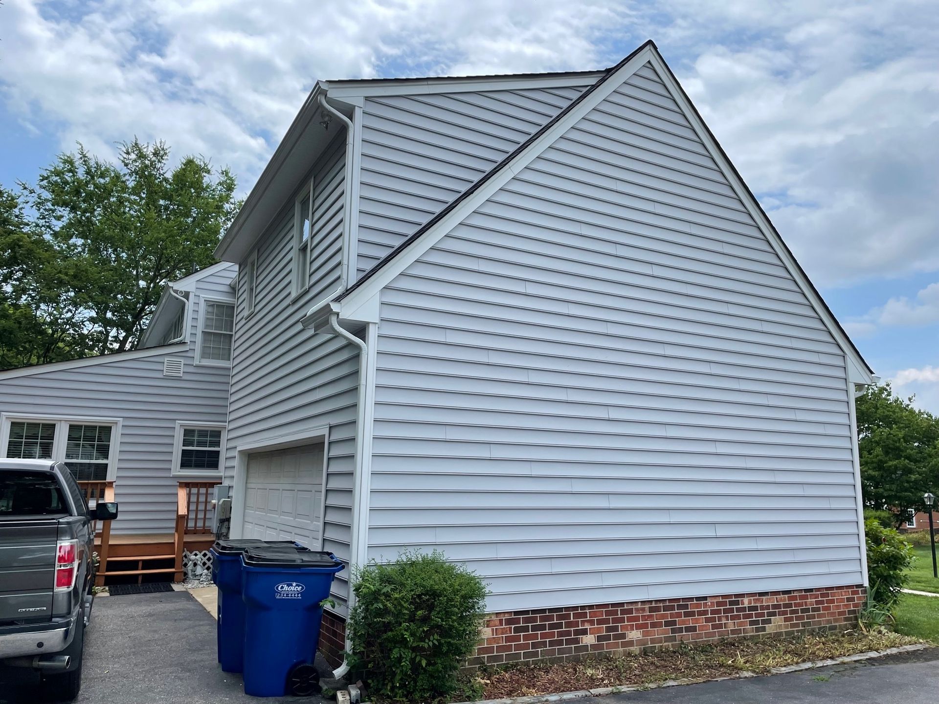 Gray-sided house exterior with brick foundation, parked truck, and blue recycling bin under a cloudy sky
