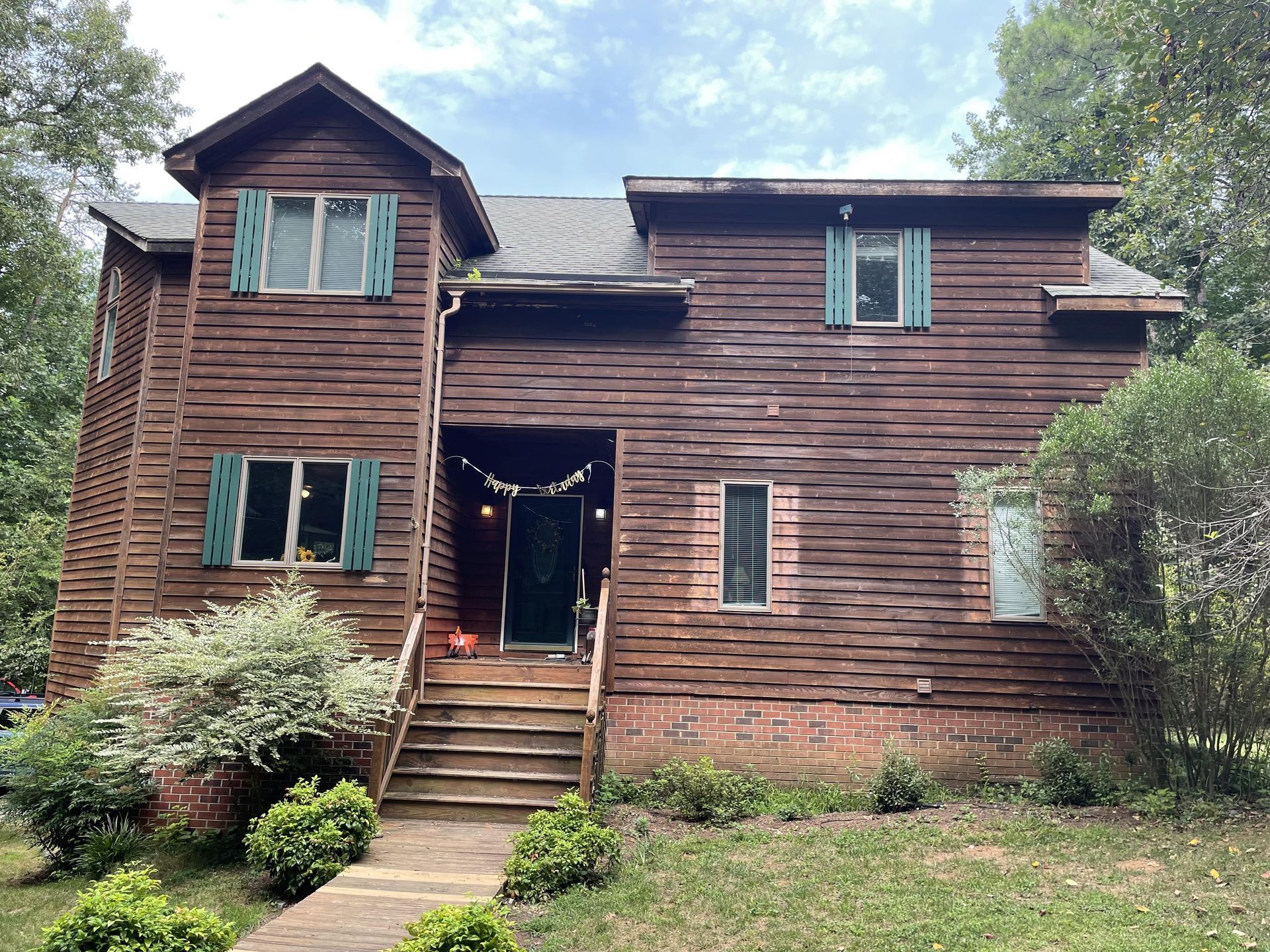 Weathered wooden house with a central front door, steps, and multiple windows amid greenery.