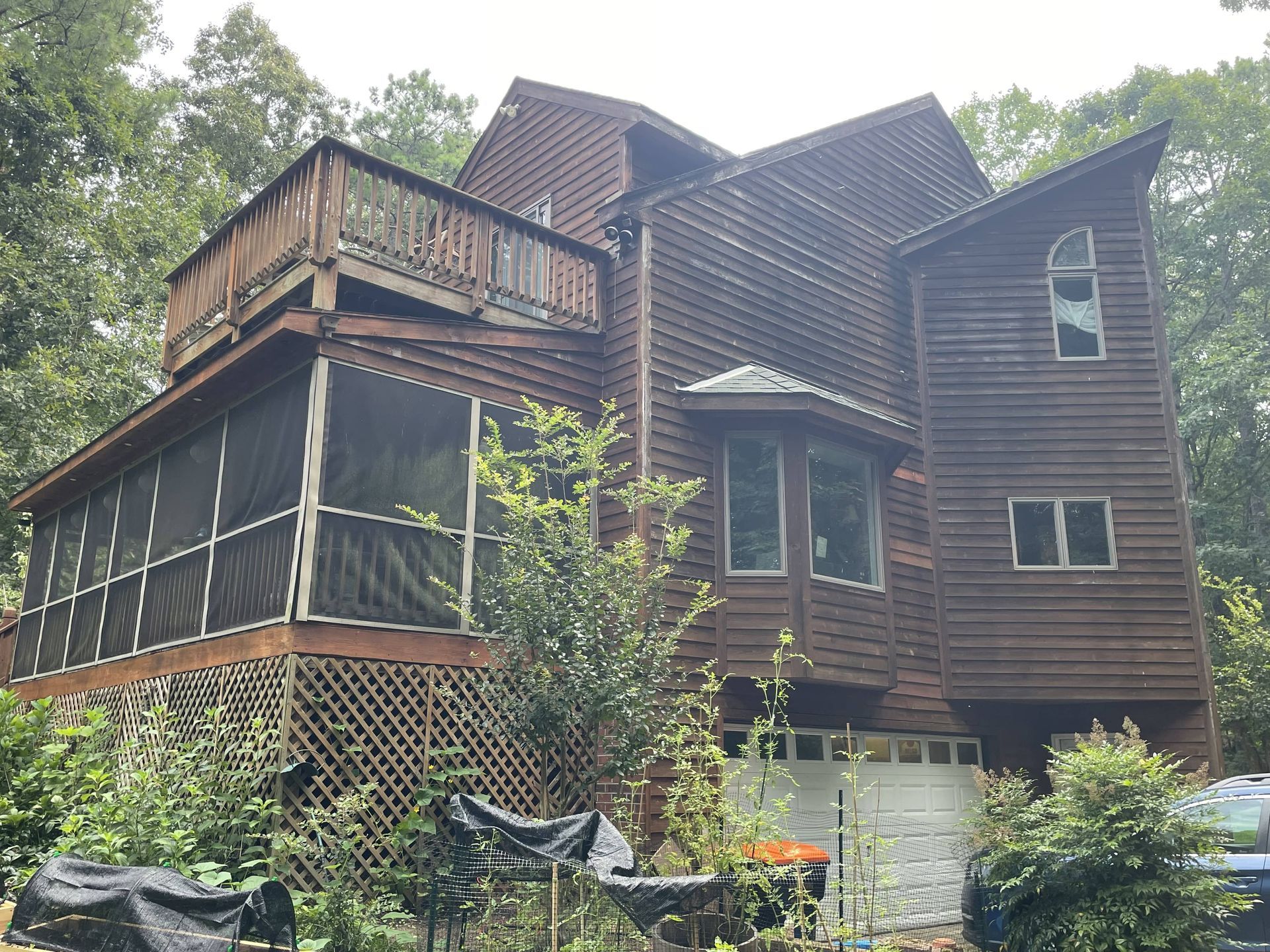 Large weathered brown house with screened porch and stone foundation, surrounded by trees and greenery