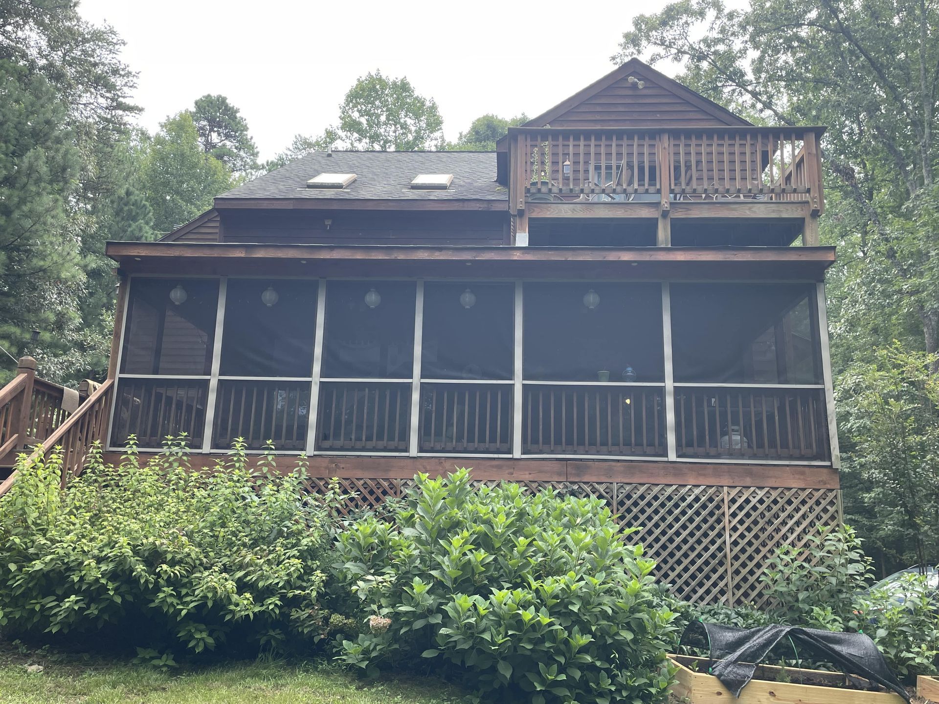 Weathered two-story cabin with screened porch, deck railing, and dense green trees and shrubs surrounding it