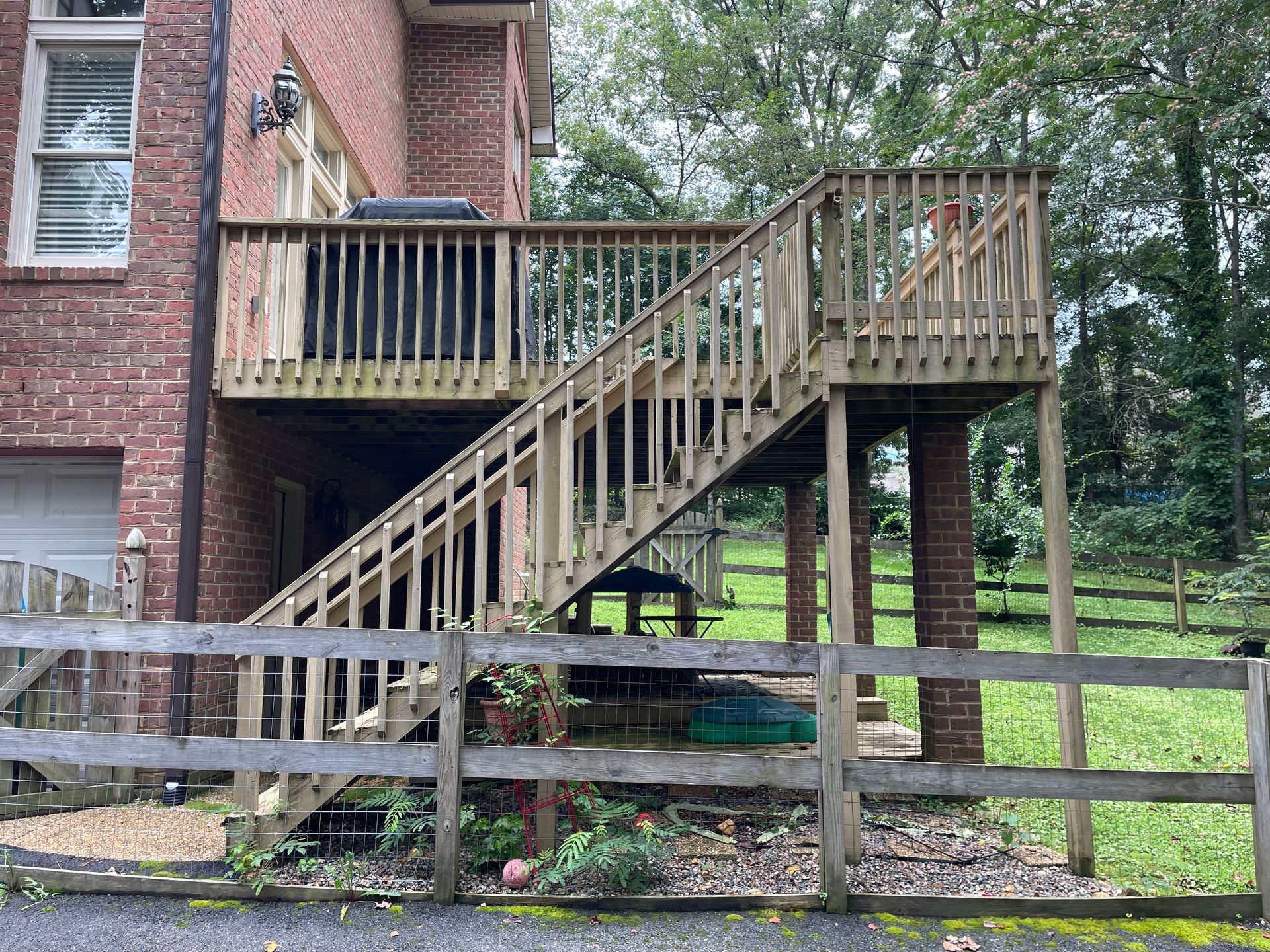 Wooden deck and stairs attached to a brick house, with a fenced yard and trees in the background.