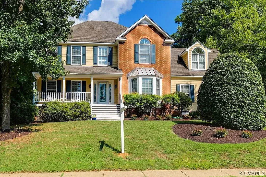 Two-story suburban house with tan siding, white trim, front porch, and landscaped lawn