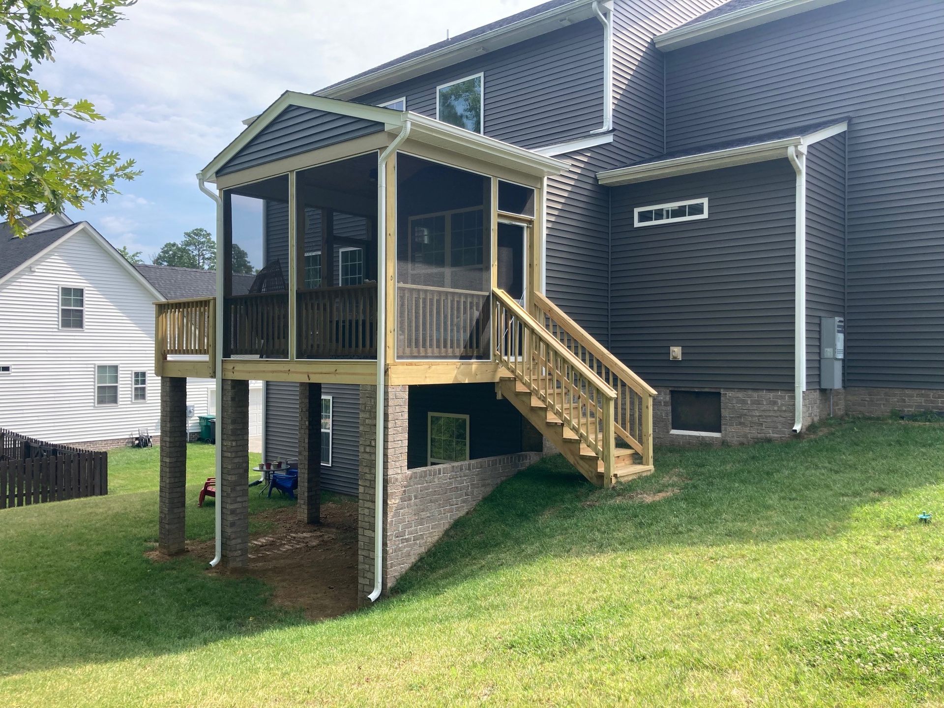 Raised back porch deck with stairs and railing attached to a gray house on a grassy slope.