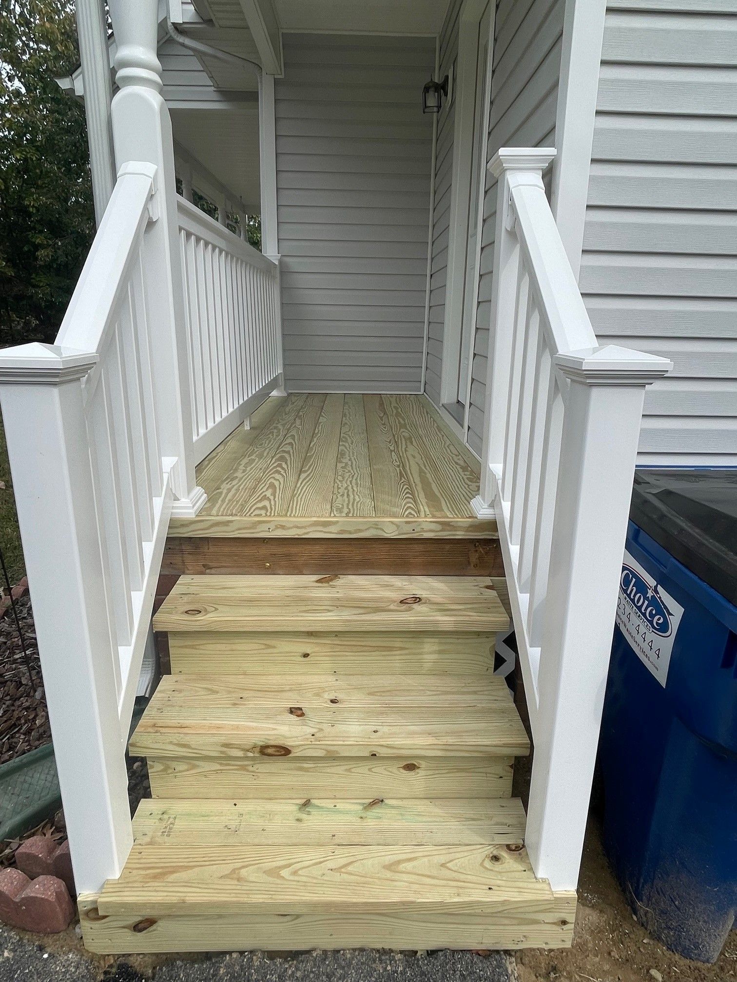 Front porch with worn wooden steps leading to a small landing, white railings, and a blue trash bin on the right