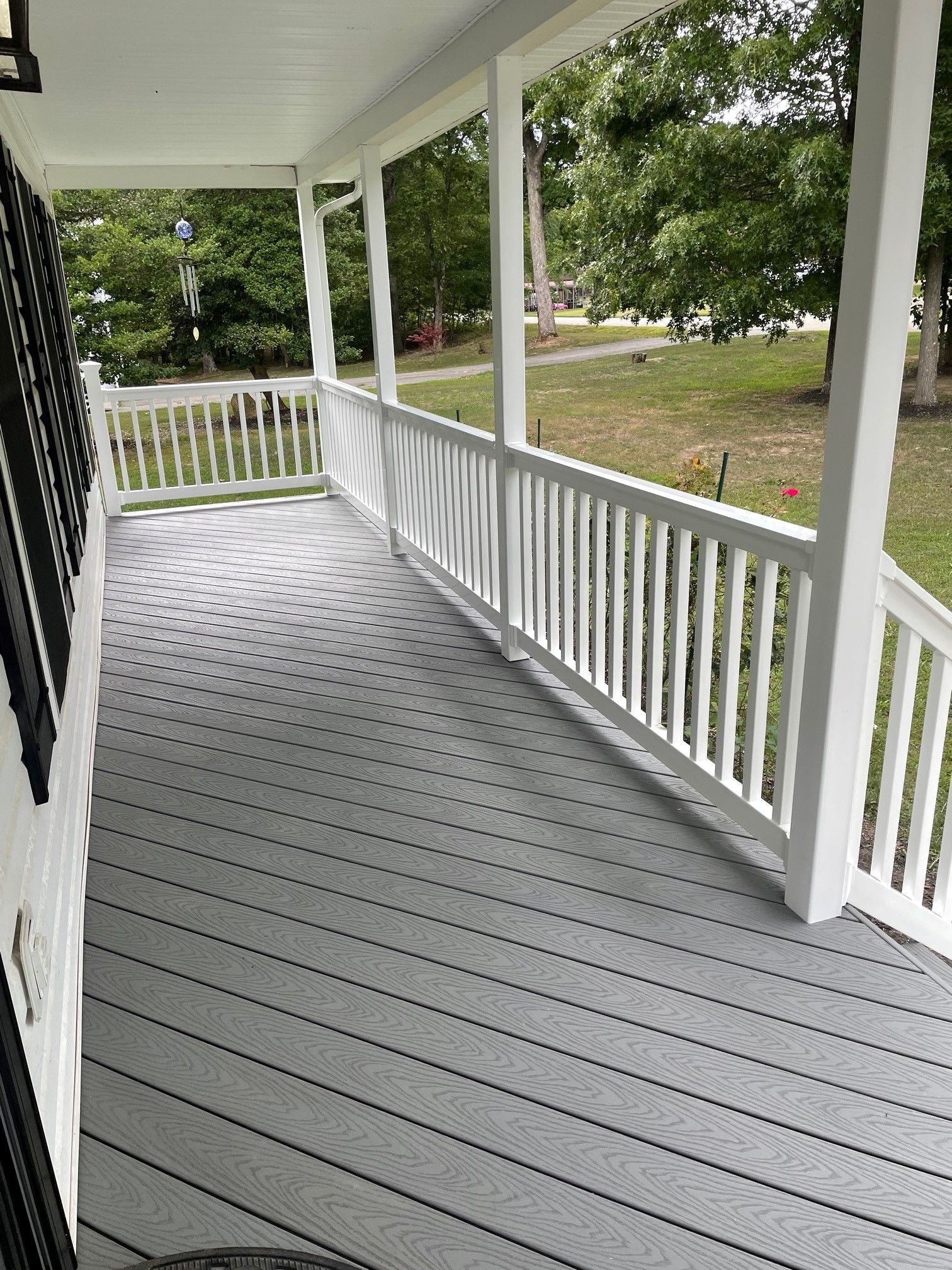 Covered porch with gray floorboards, white railings, and a view of green trees beyond.