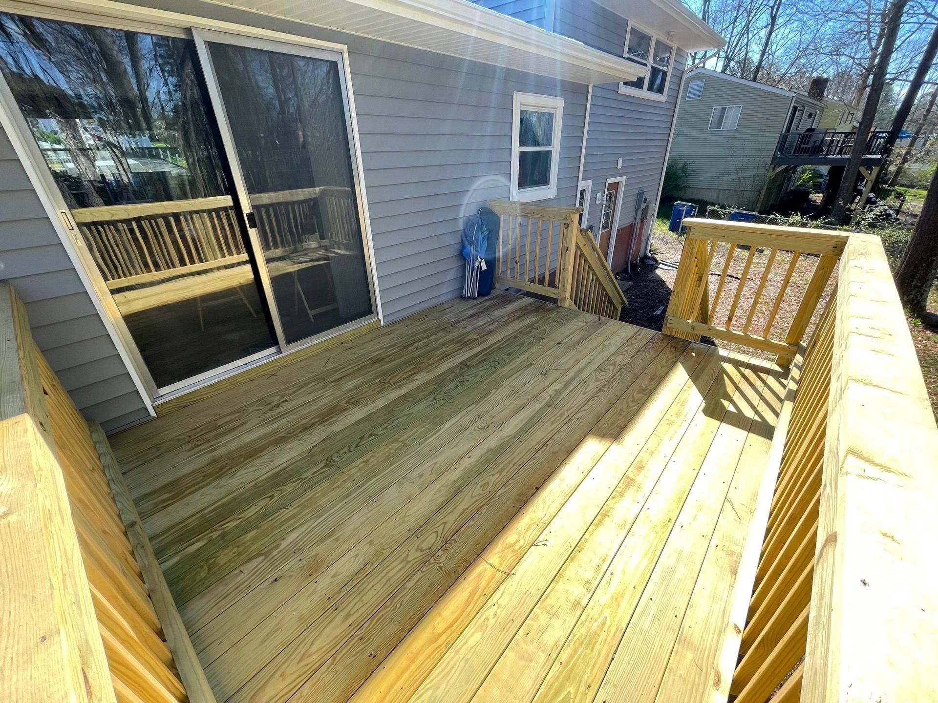 Freshly built wooden deck beside a gray house, with sliding glass door and stairs railing in sunlight