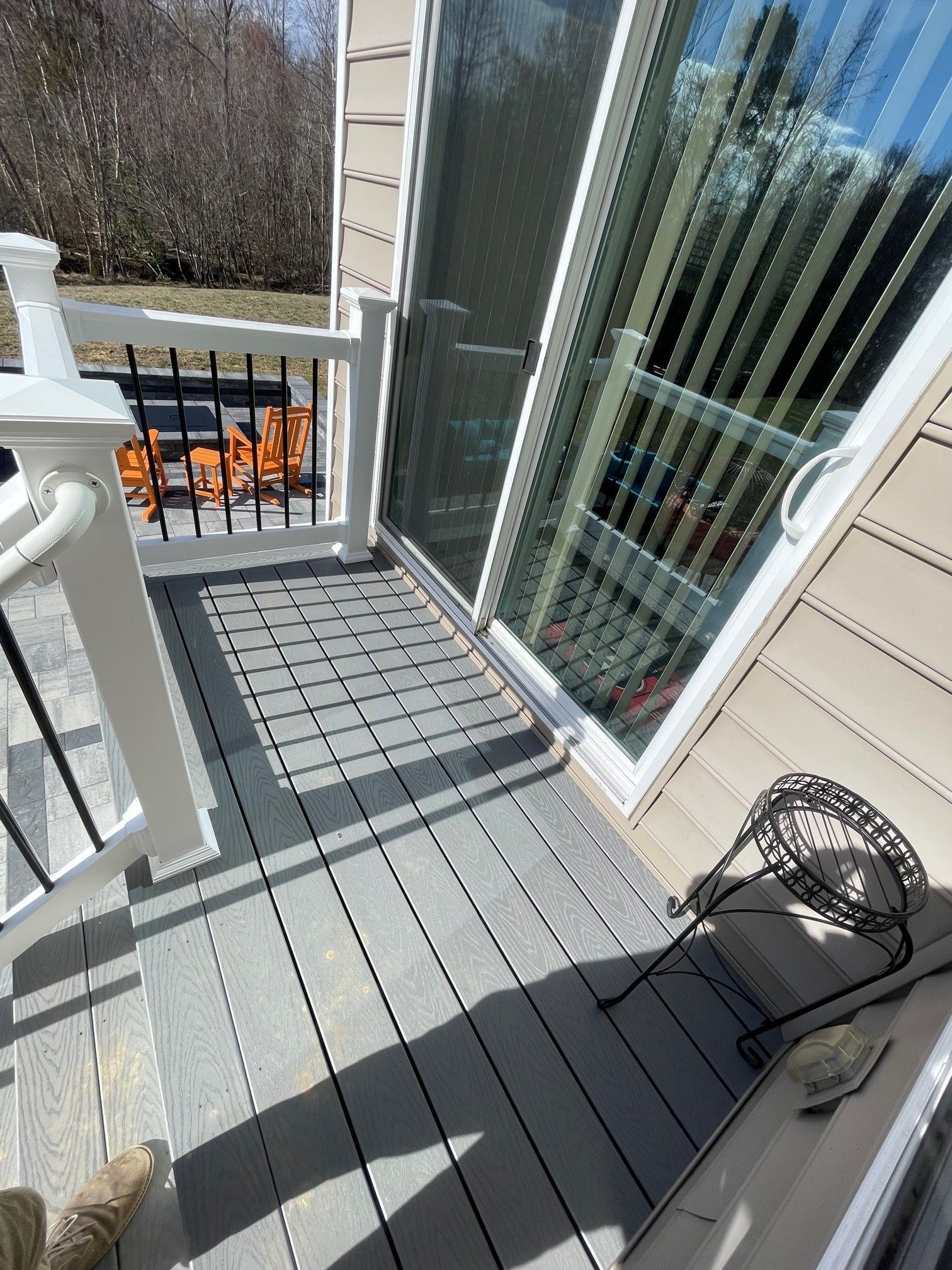 Small porch with gray deck boards, sliding glass door, white railing, and a black wire side table in sunlight.