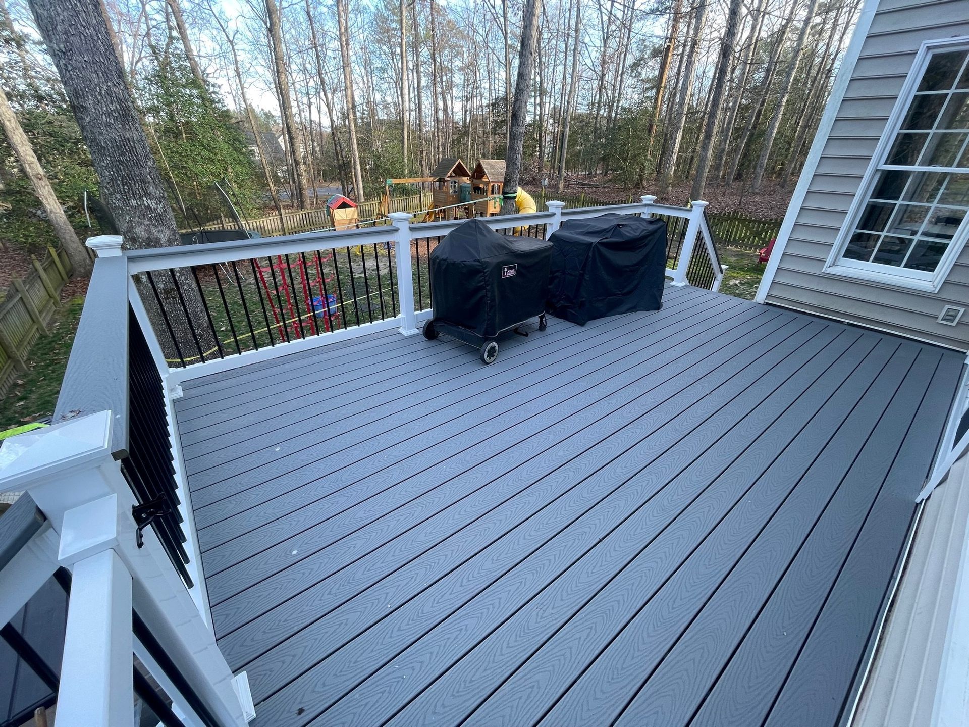 Gray deck with black grill cover, railing, and wooded backyard view beside a house siding