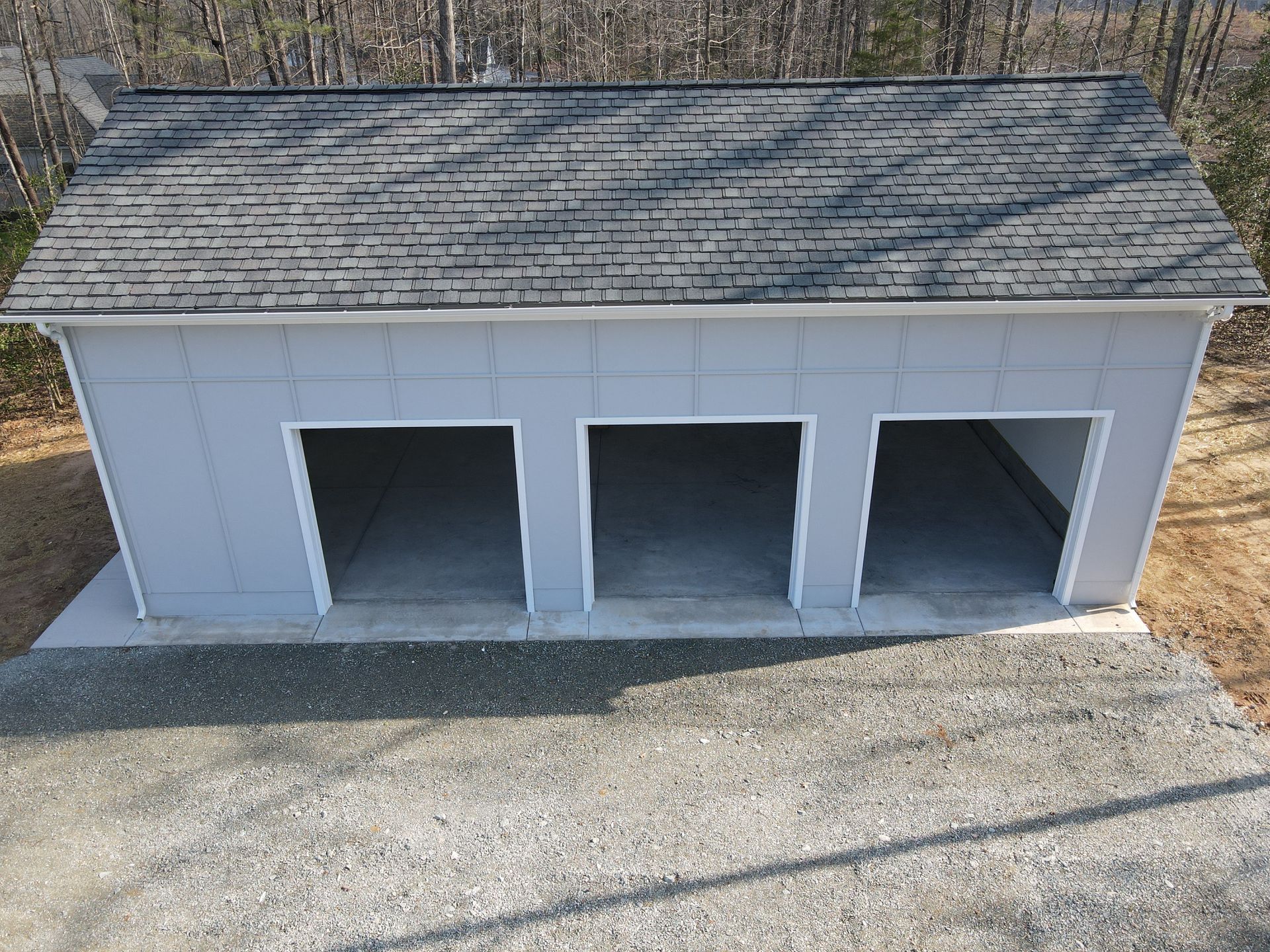 White three-bay garage or shed with gray shingled roof on a gravel lot outdoors.