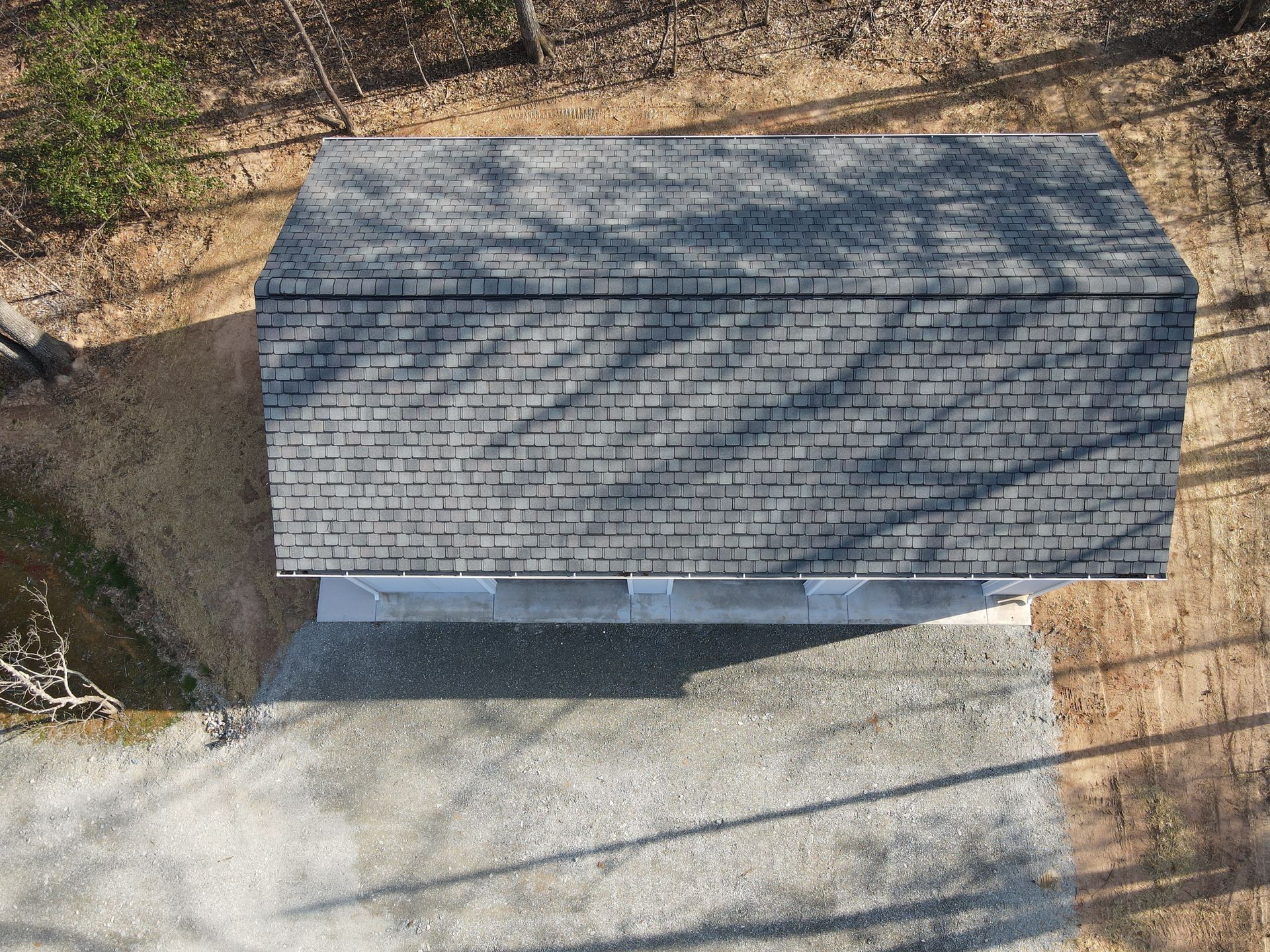 Aerial view of a small gray-roofed building beside a paved area and trees.