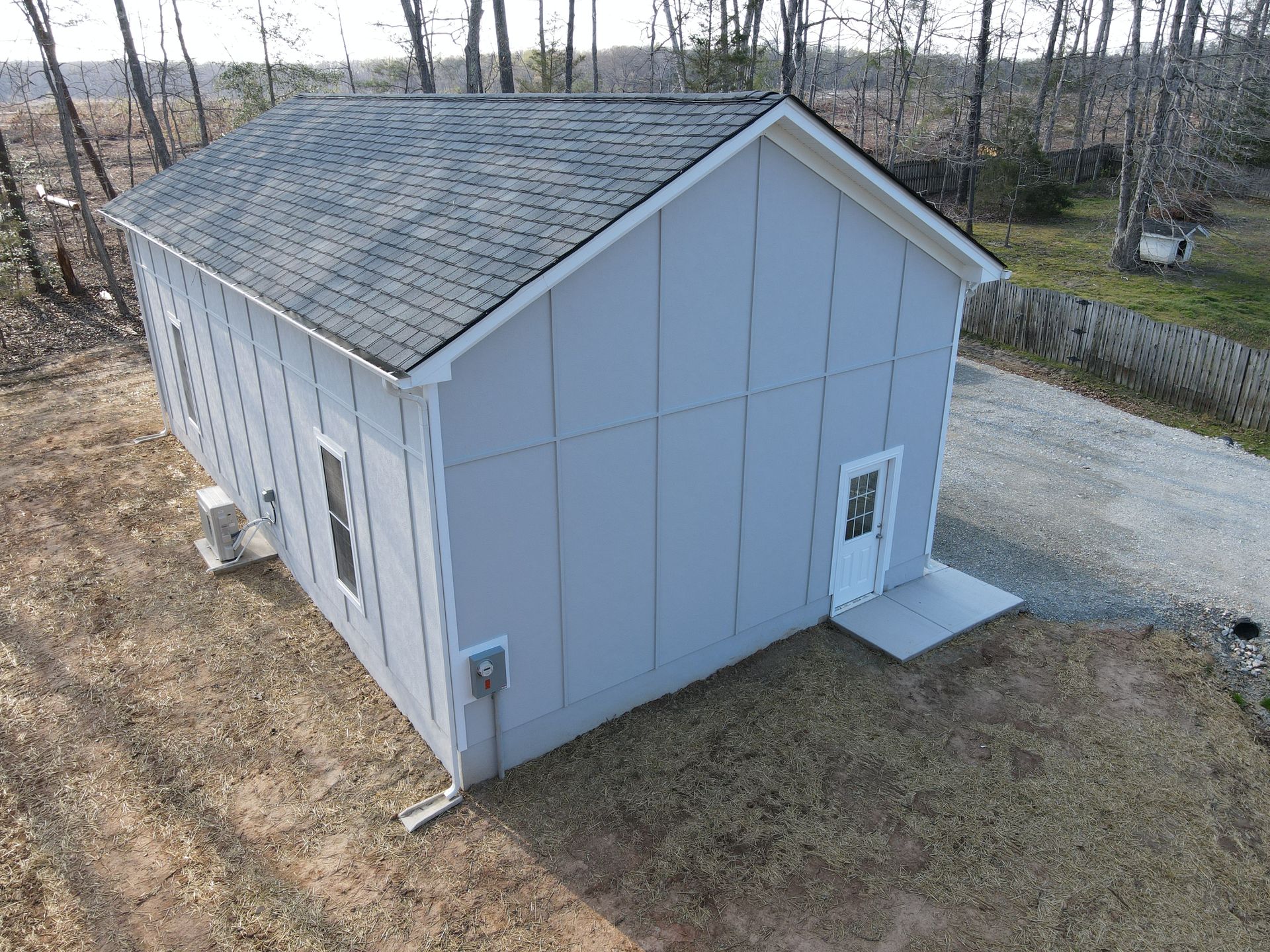 Small gray shed with a gabled roof on a gravel lot, surrounded by leafless trees