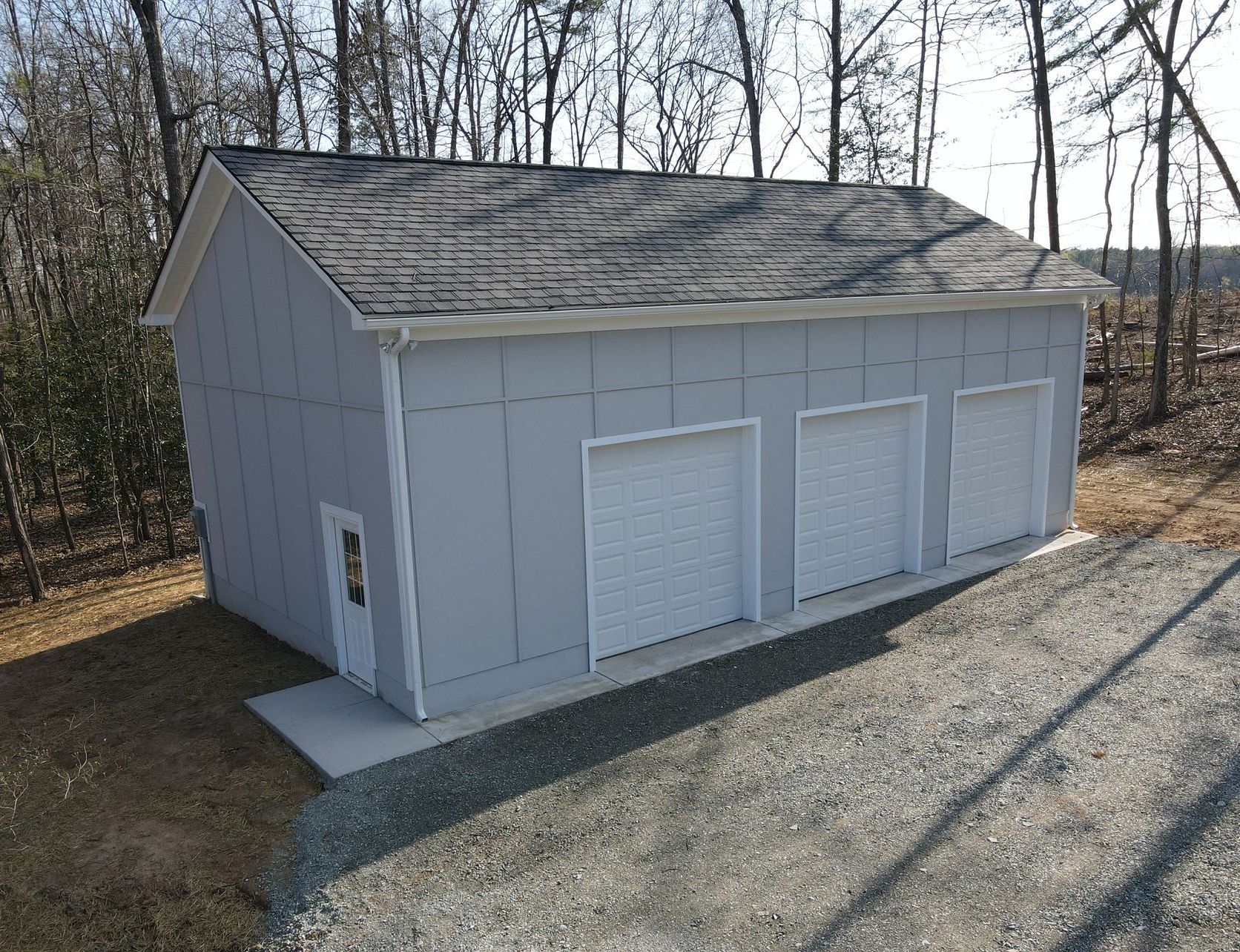 Small white garage or storage shed with three doors, set on a gravel driveway beside woods.