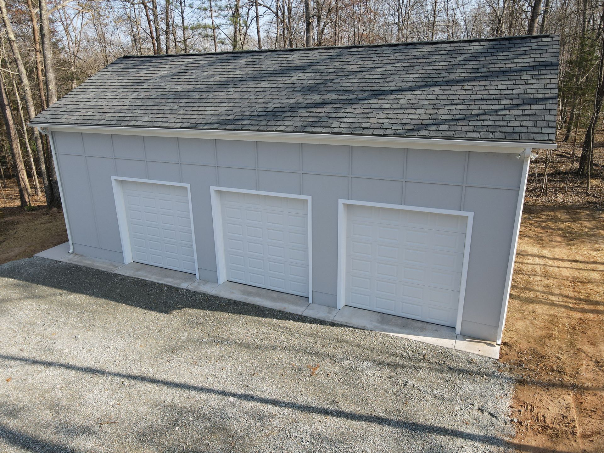 Gray detached garage with three white doors and shingled roof beside a wooded area