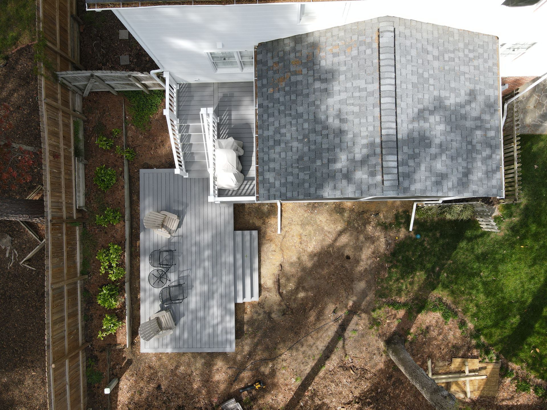 Aerial view of a house roof, backyard patio, and fenced yard with trees and dirt path.