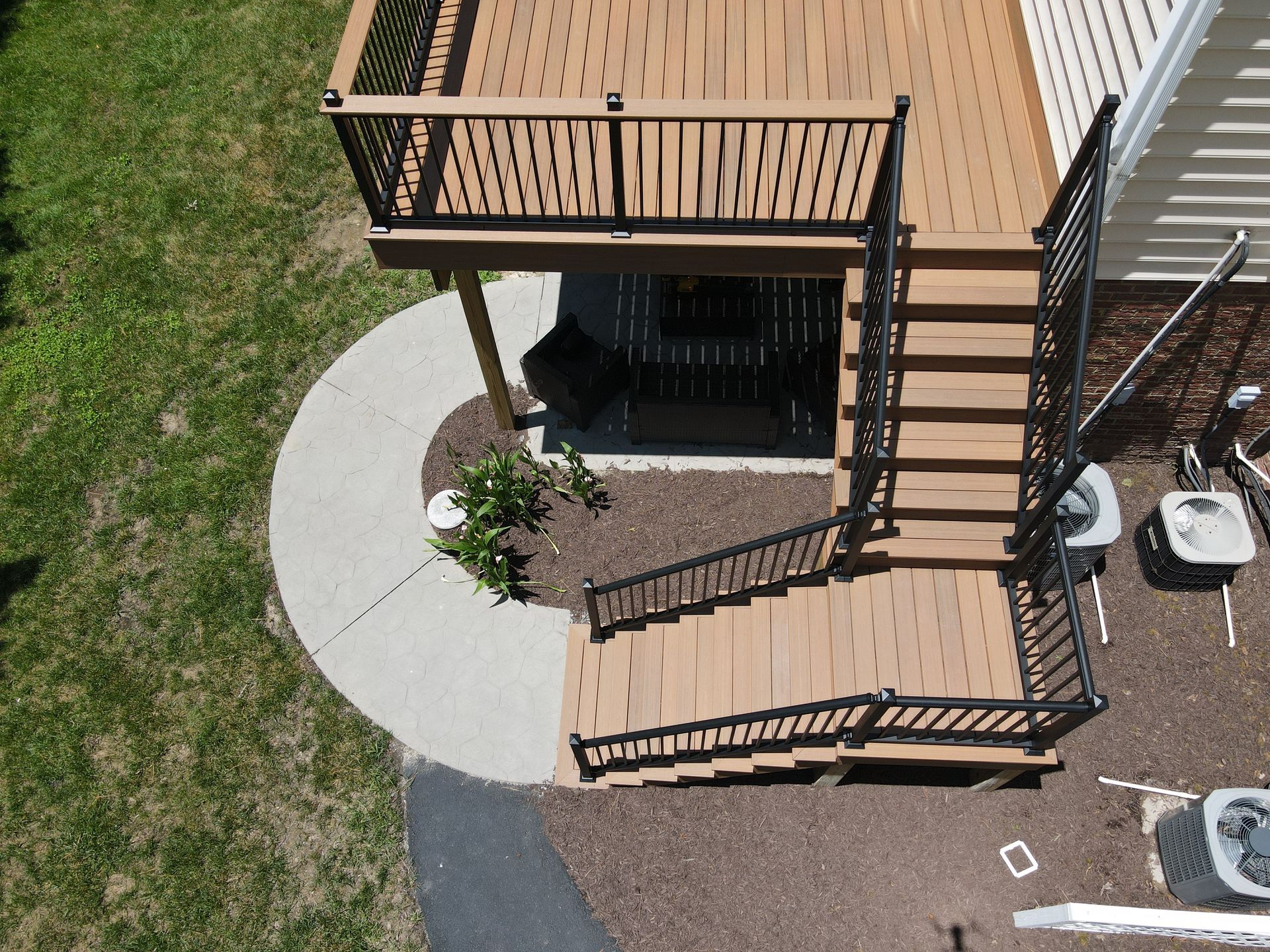 Overhead view of a wooden deck with stairs and a small patio, beside a curved concrete path and lawn