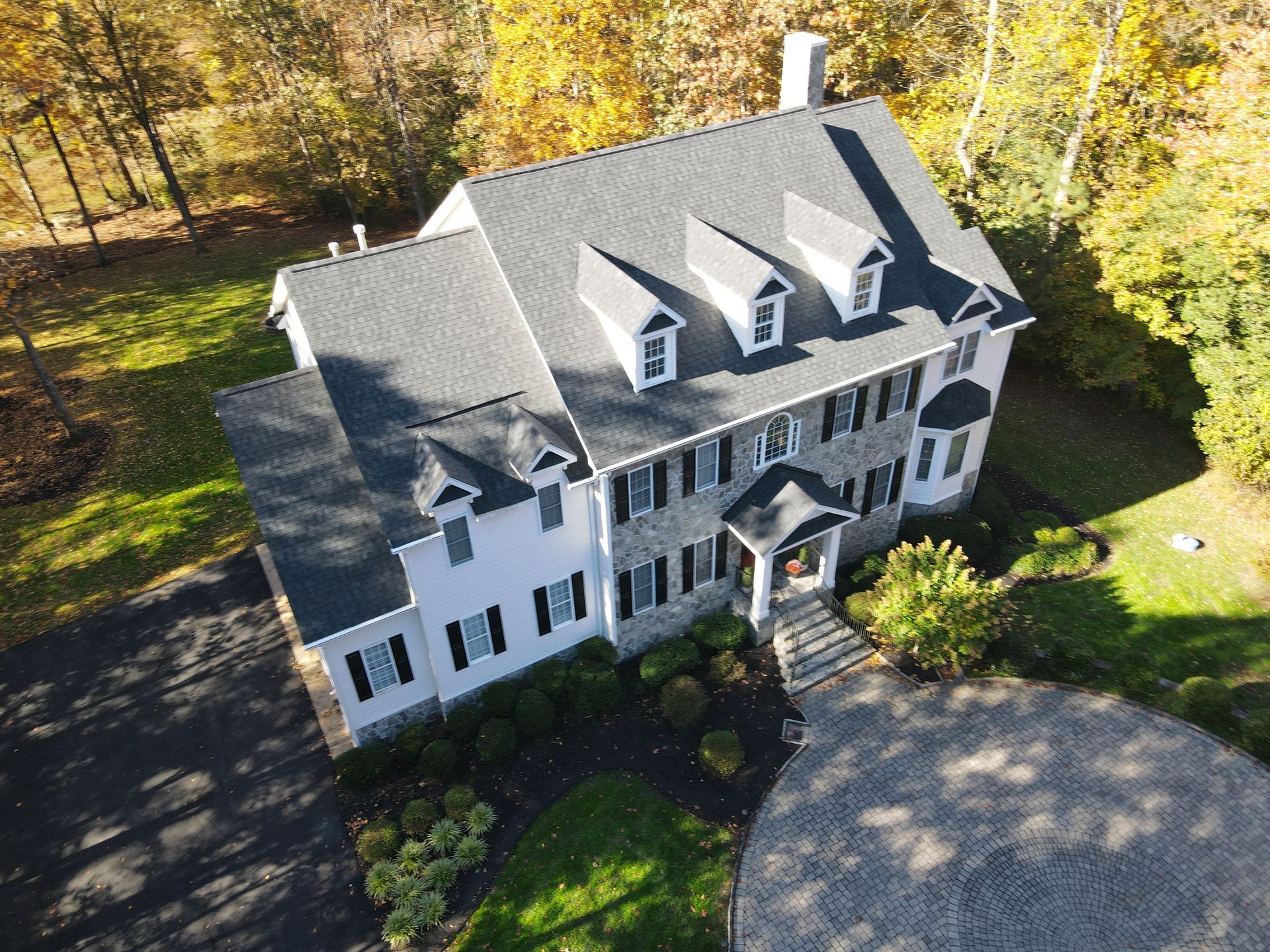Aerial view of a large gray house with a steep roof and white trim, surrounded by autumn trees.