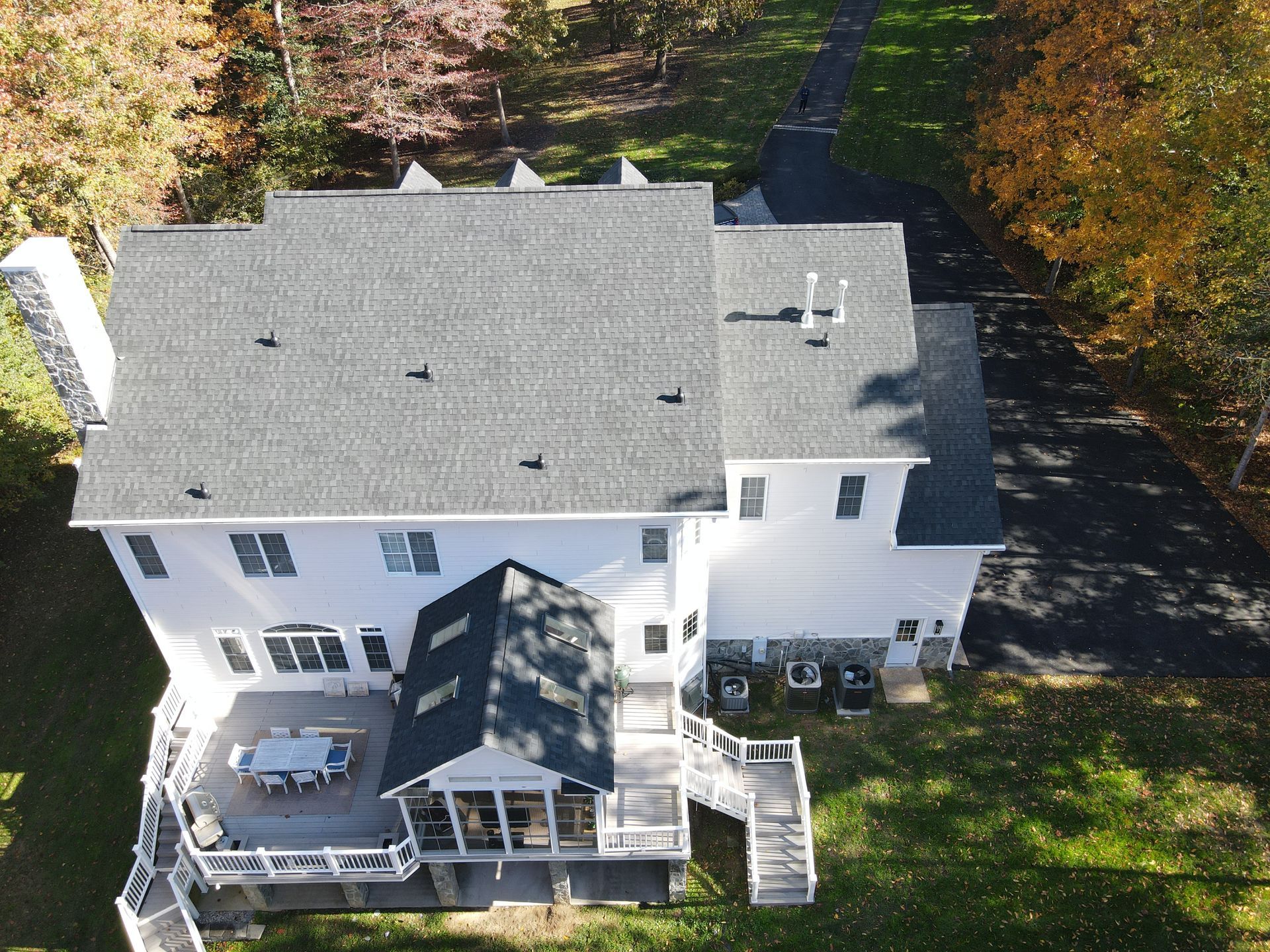 Aerial view of a white two-story house with a gray roof, deck, and surrounding trees in autumn.