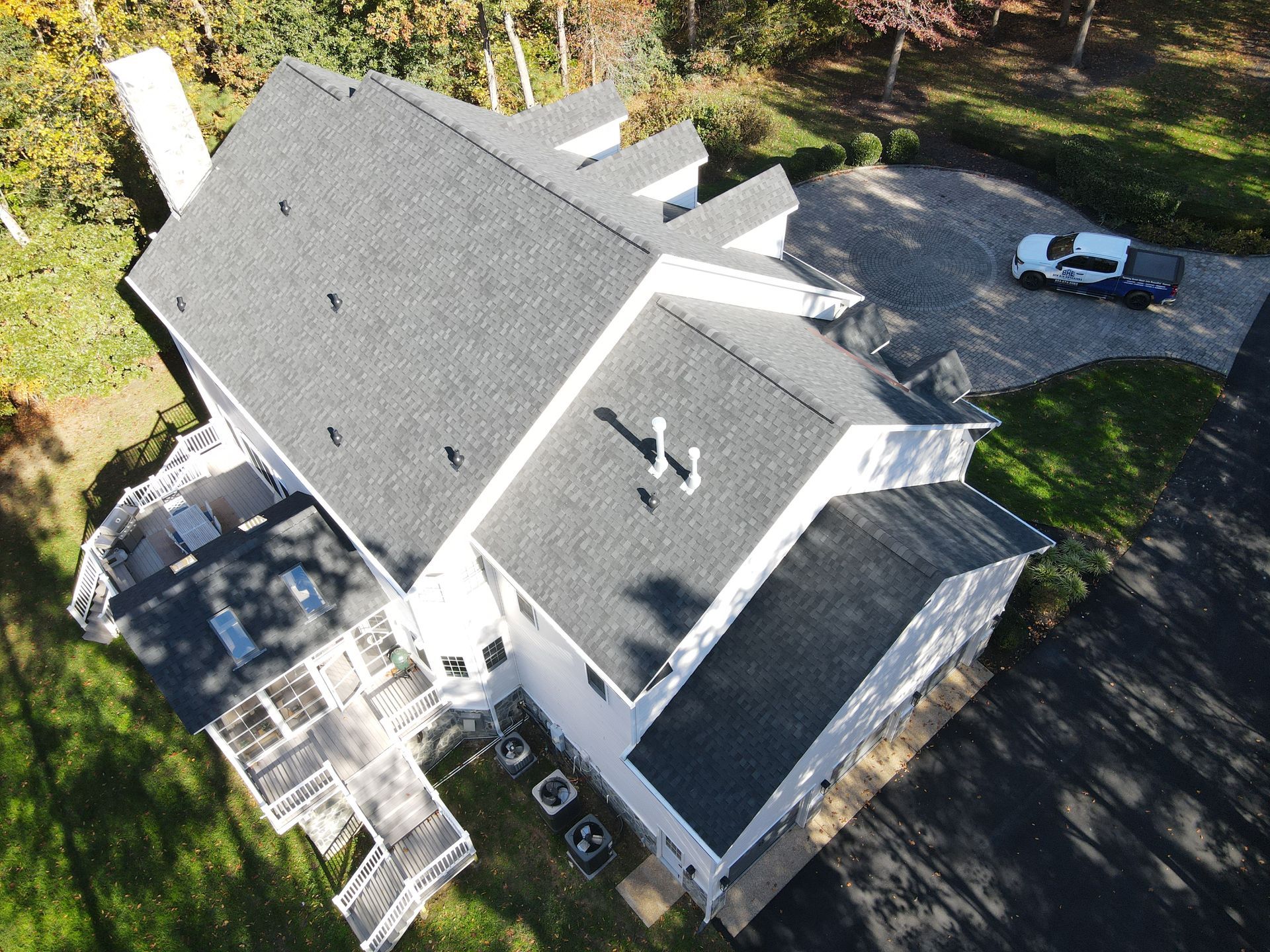 Aerial view of a white house with gray roof, deck, and parked car beside a wooded driveway.