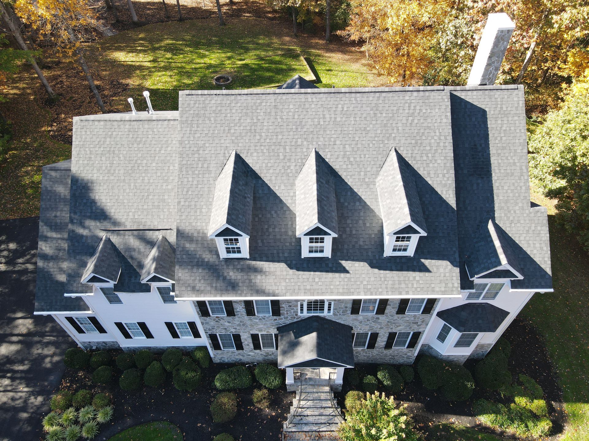 Aerial view of a gray house with a dark roof, white trim, and several dormer windows surrounded by trees.