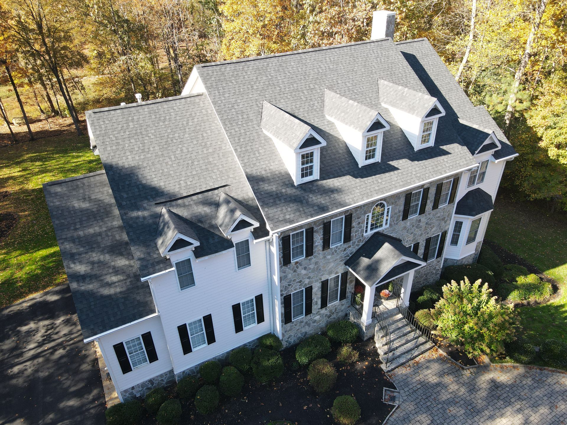 Aerial view of a large gray-and-white house with dormer windows, surrounded by autumn trees.