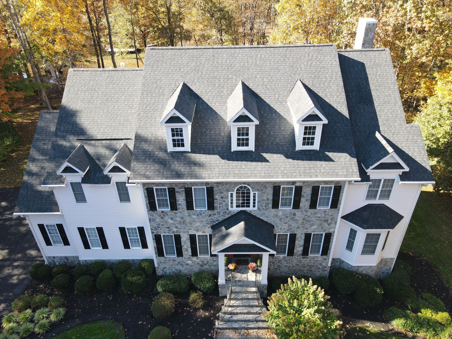 Aerial view of a large white-and-gray house with dormer windows, stone facade, and autumn trees behind it