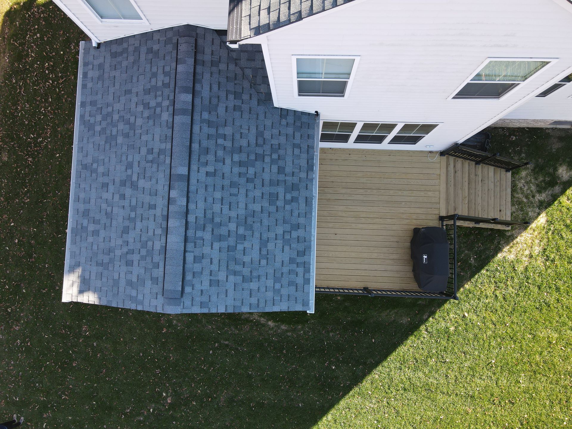 Aerial view of a house roof, deck, and green lawn with a trash bin beside the deck.