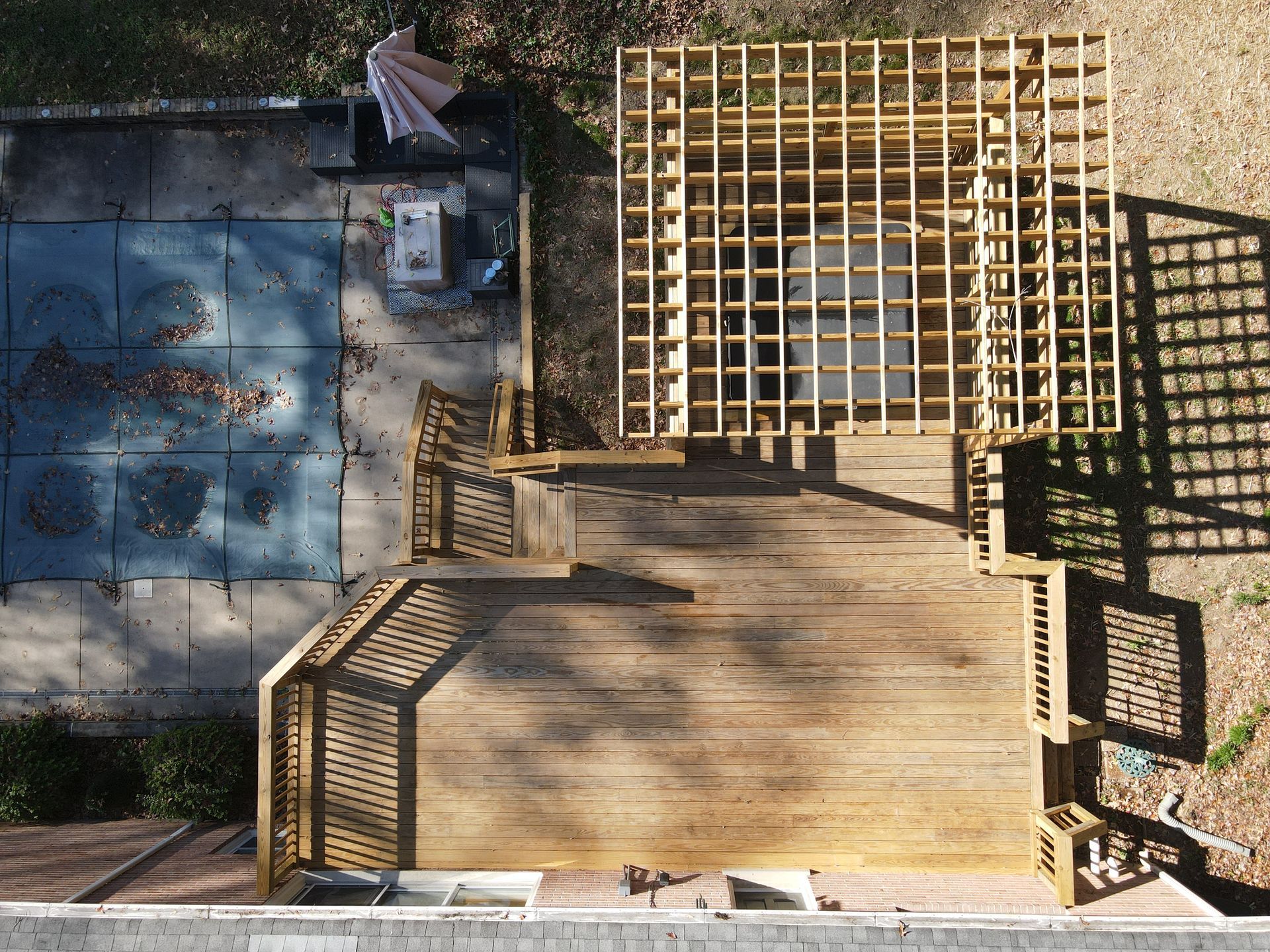 Top-down view of a backyard deck and patio beside a blue pool, with lattice fencing and strong sunlight shadows