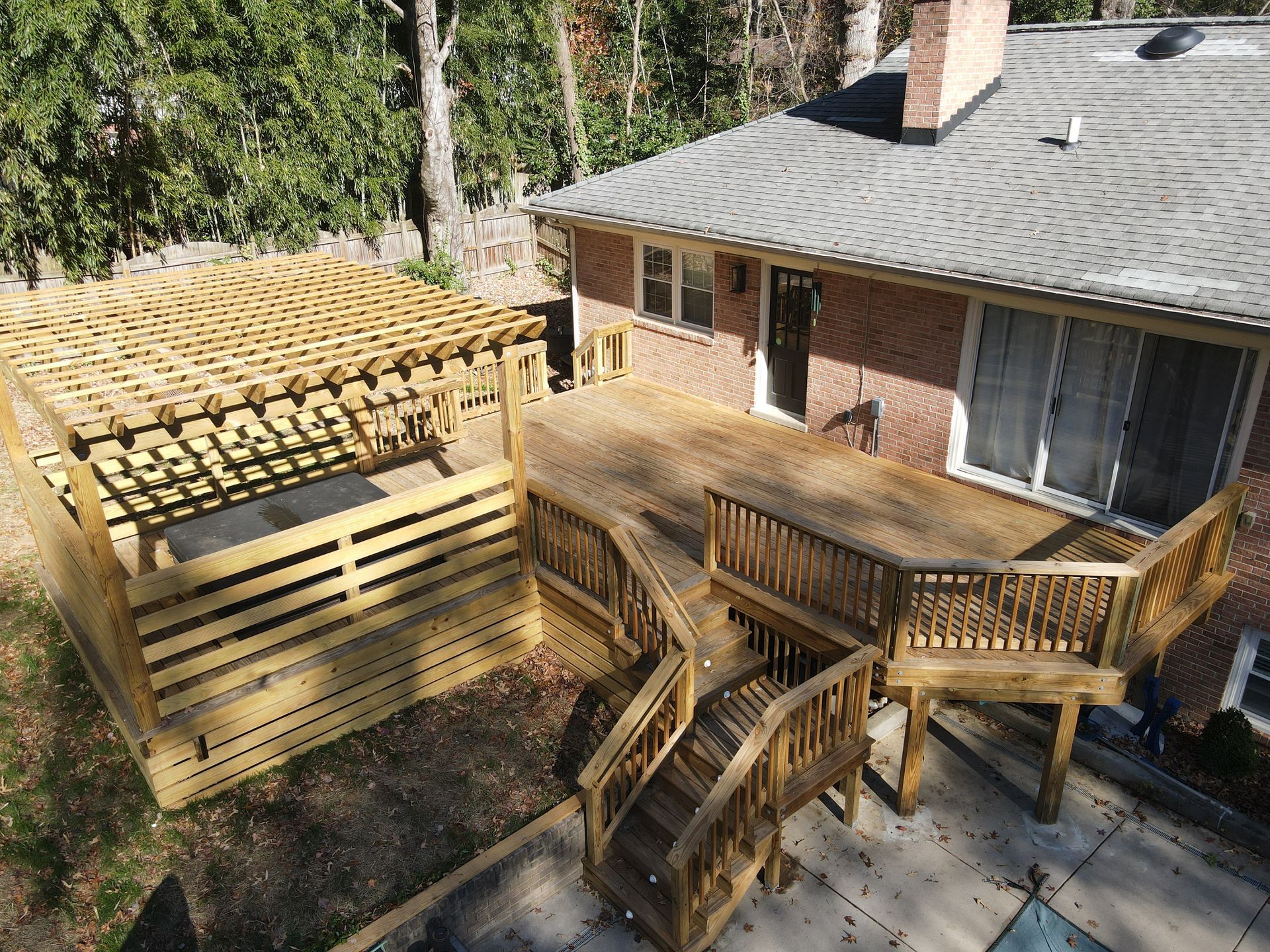 Backyard deck with wooden railing and stairs beside a brick house, shaded by tall trees