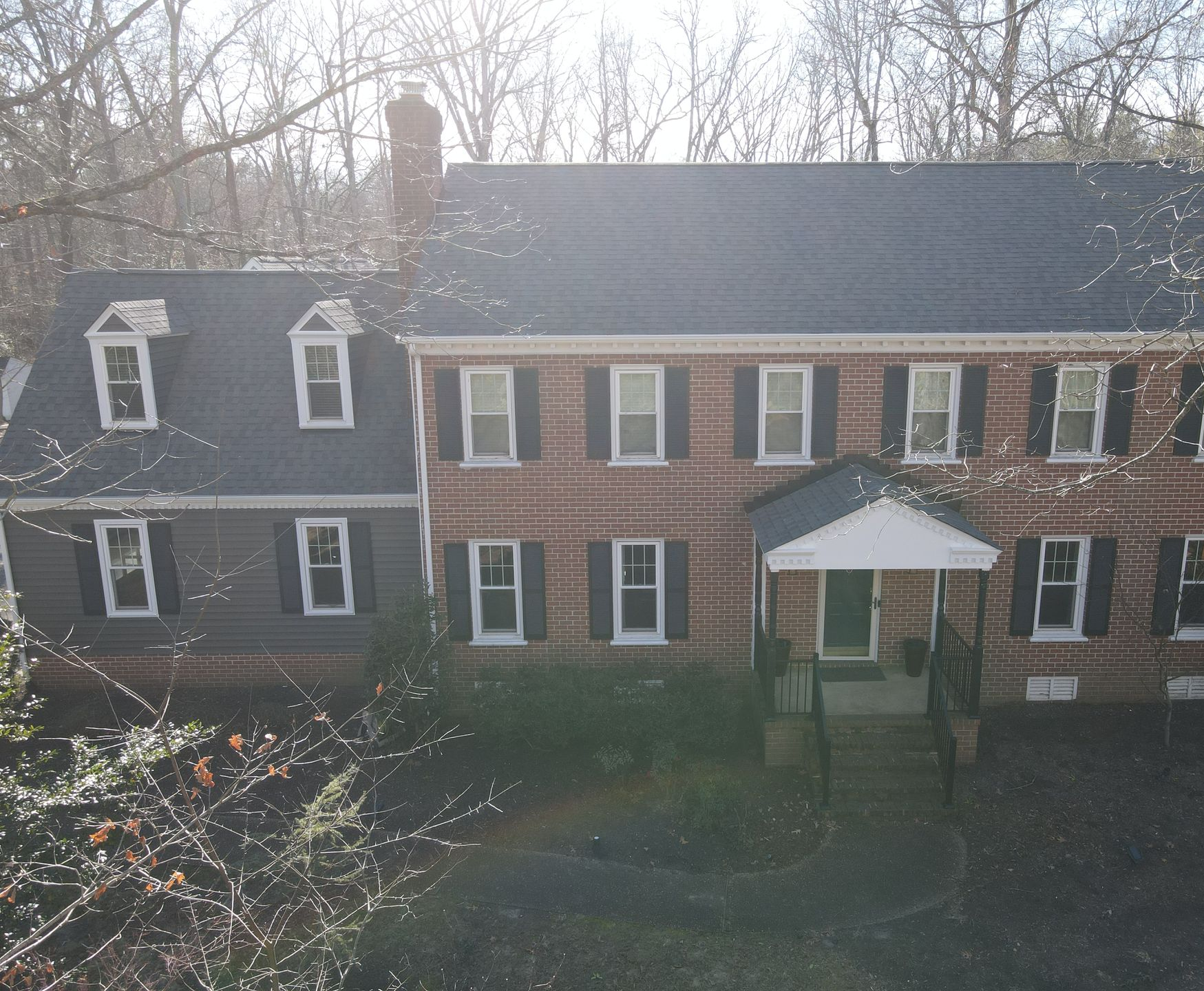 Large brick and white house with dormer windows and a covered front porch in a wooded yard