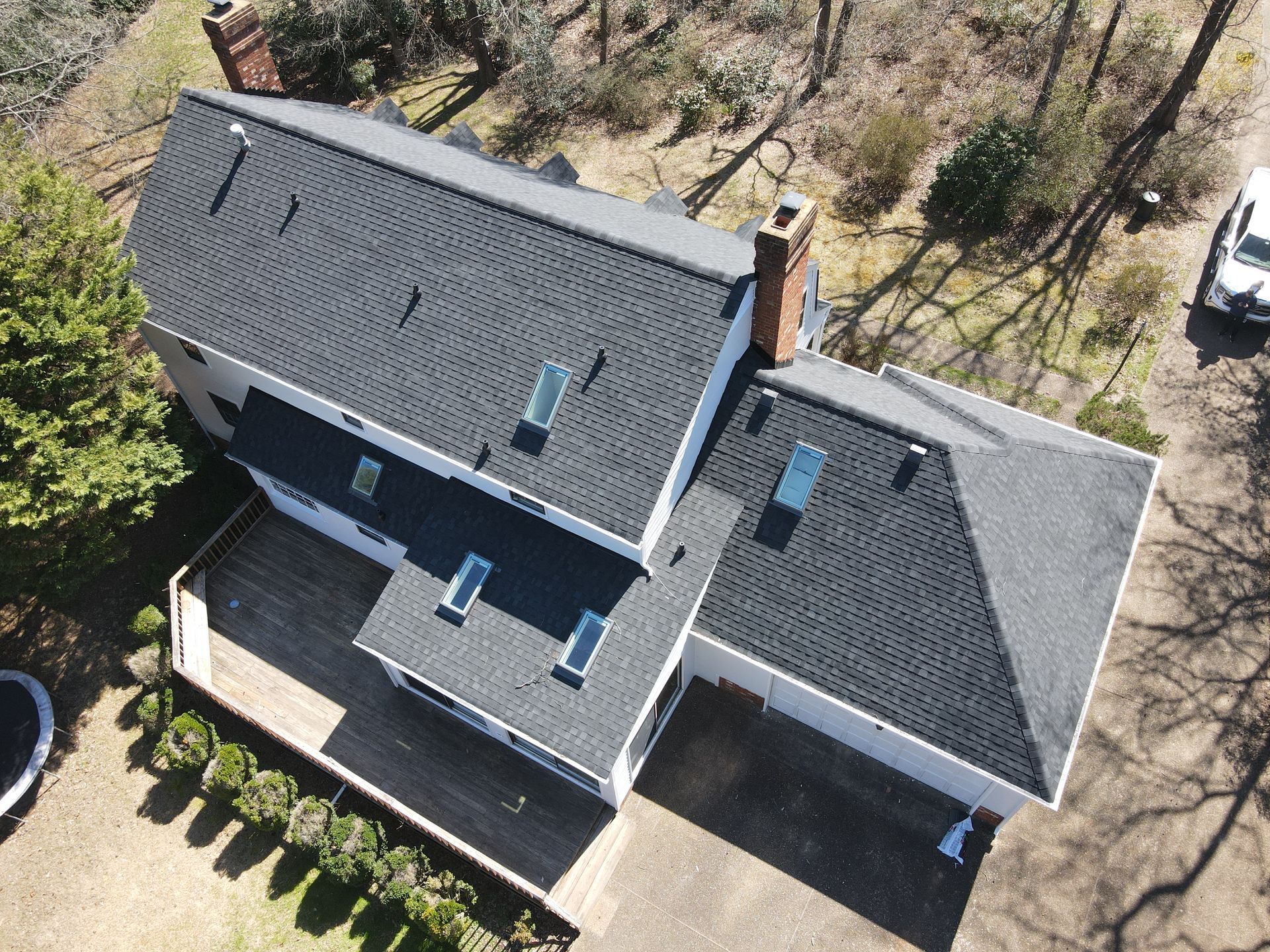 Aerial view of a gray-roofed house with multiple skylights and a deck, surrounded by trees.