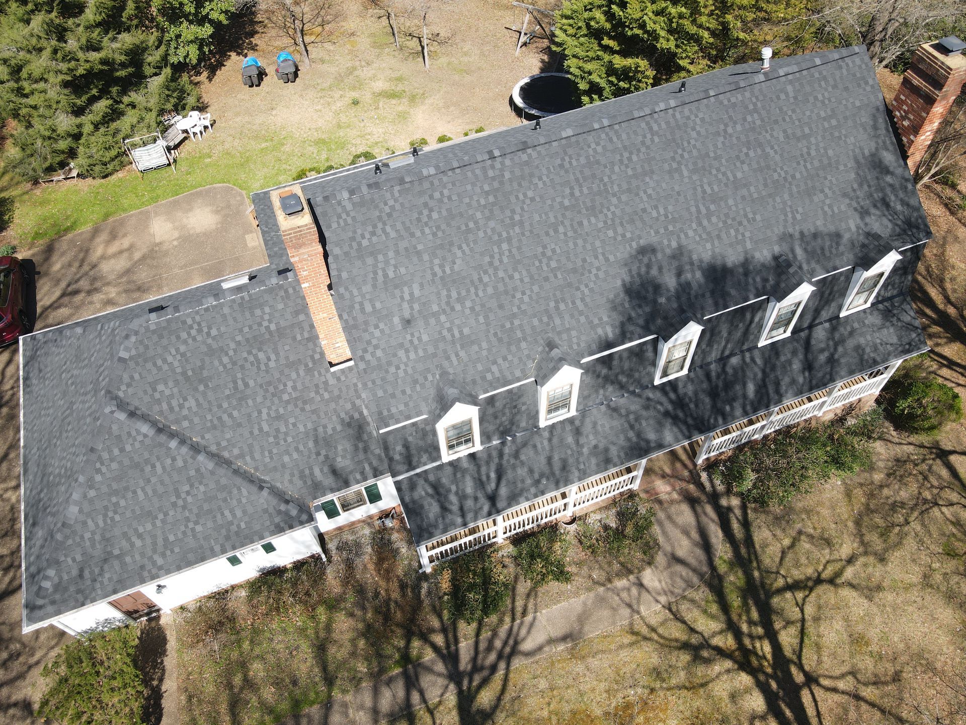 Aerial view of a gray-shingled house roof with chimneys, dormer windows, and trees around it