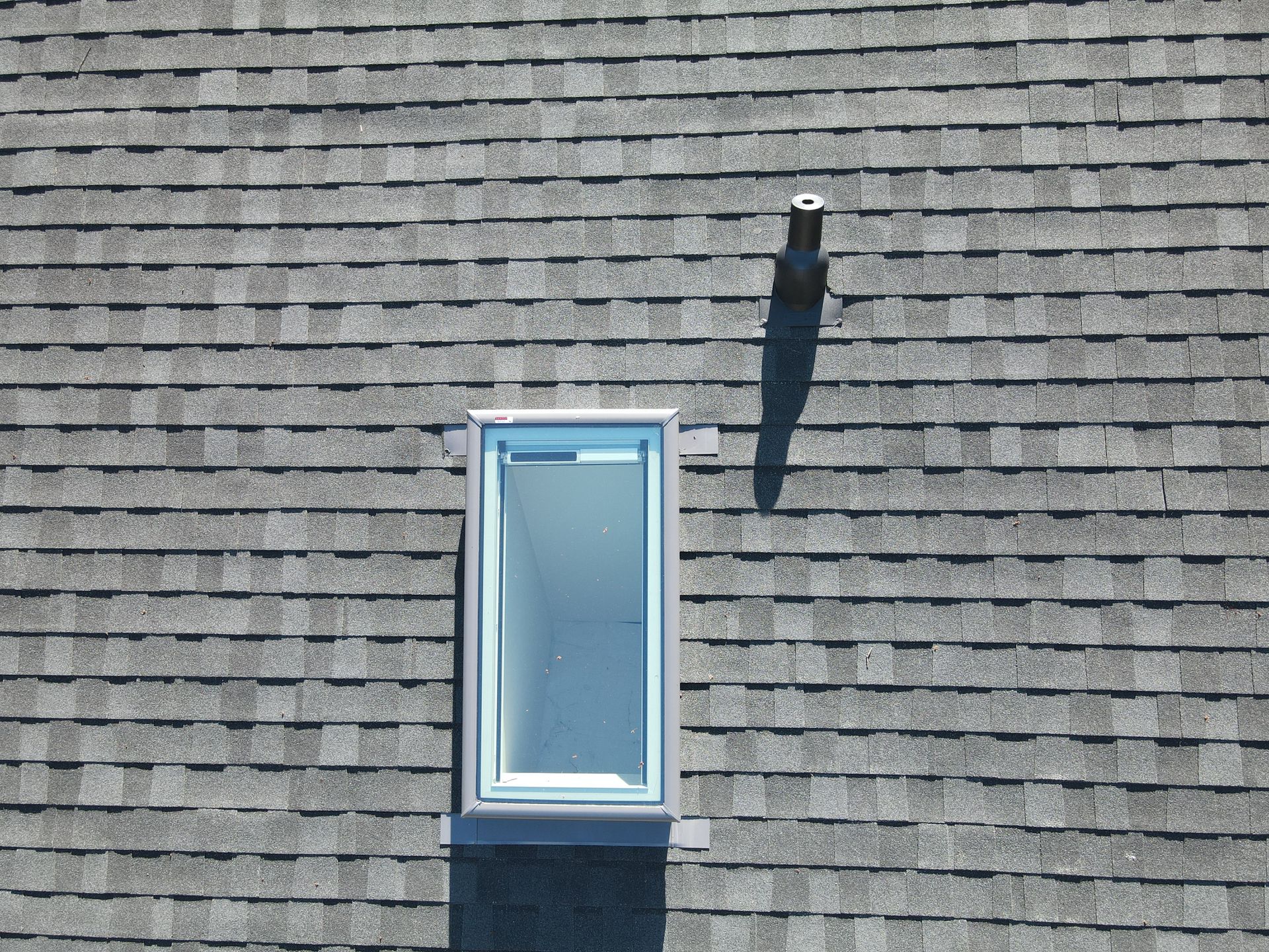 Small blue-framed window in gray shingled roof with a vent pipe nearby