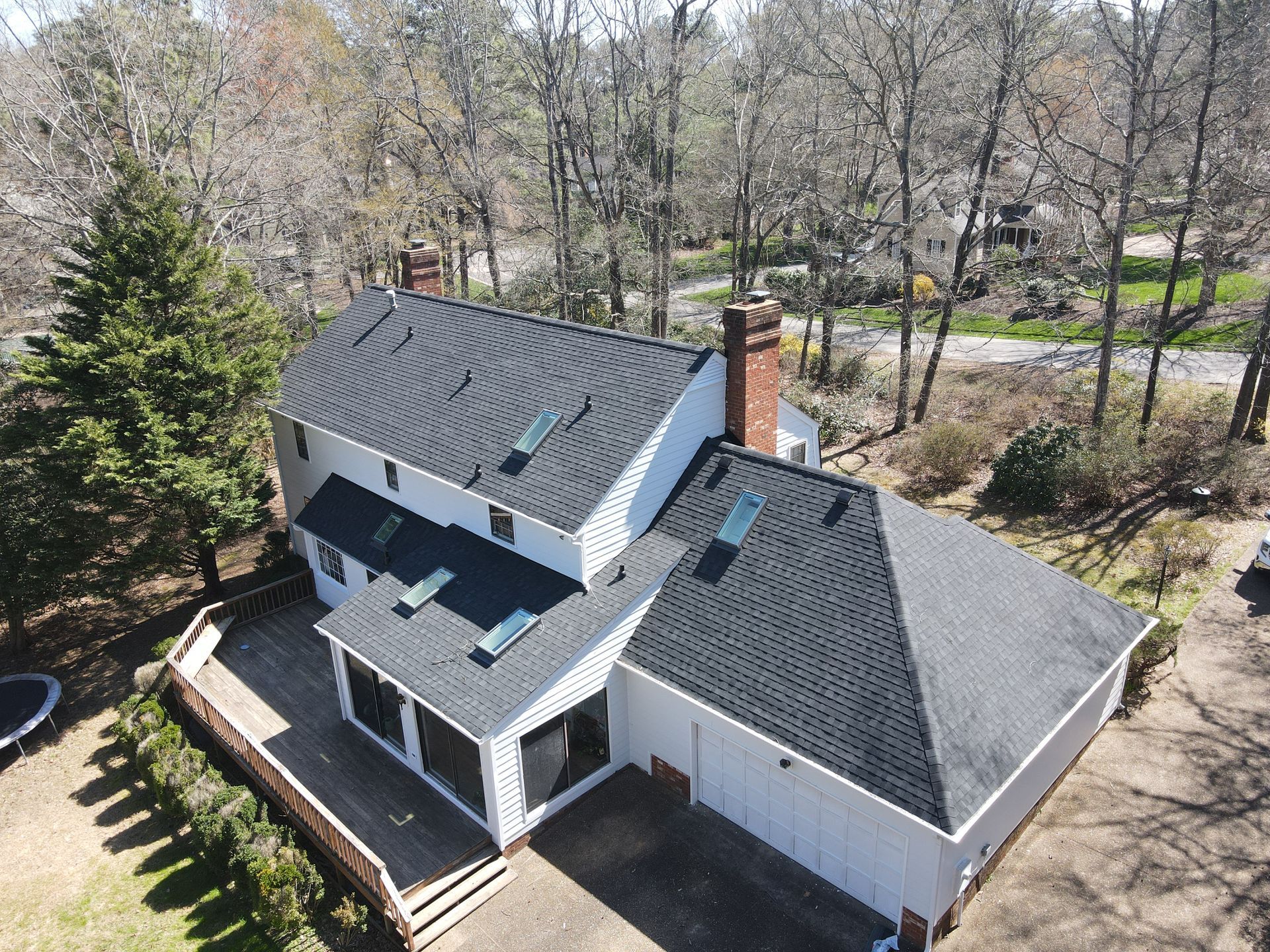 Aerial view of a gray-roofed house with a deck, brick chimney, and surrounding trees.