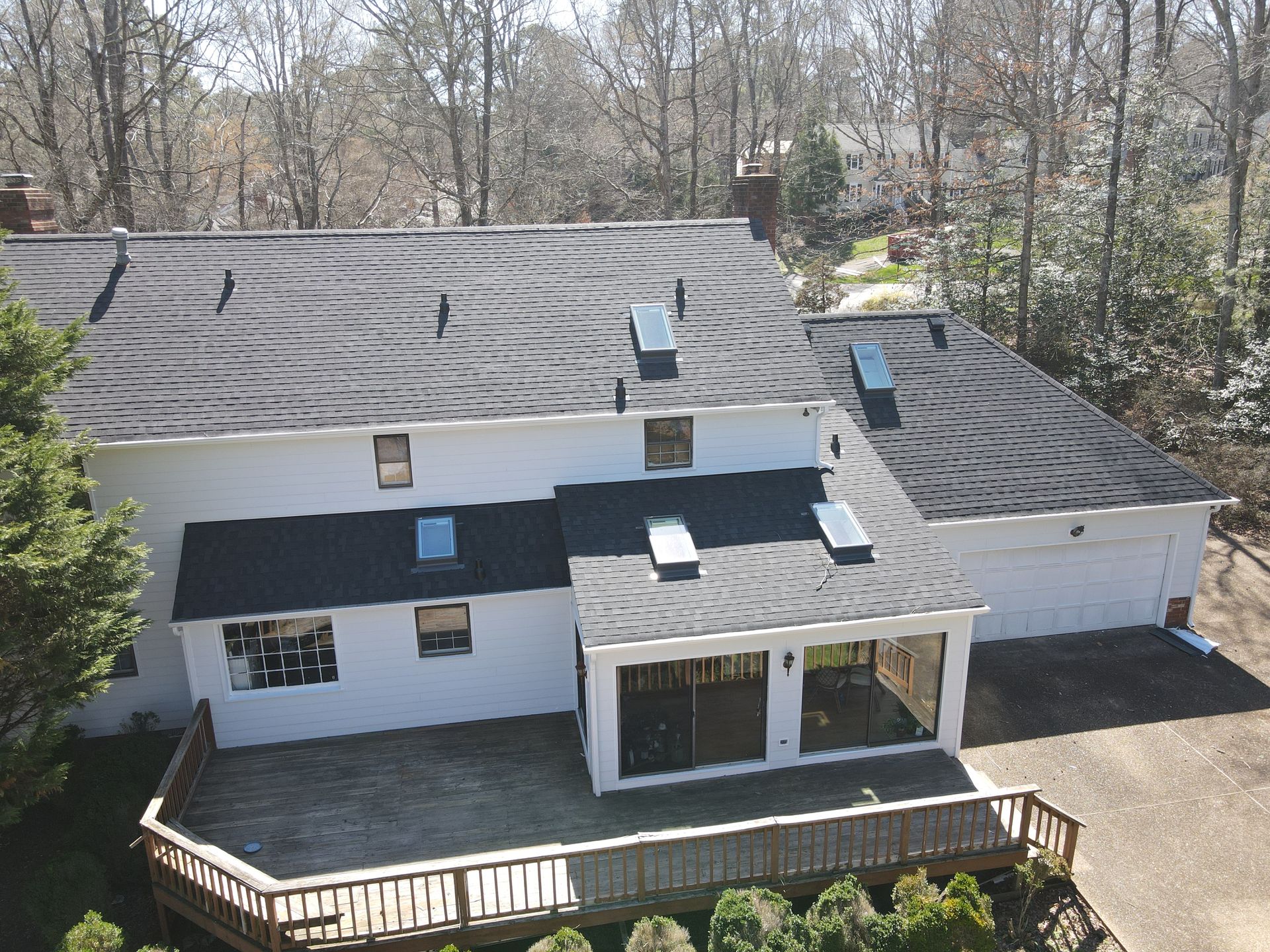 Aerial view of a white house with a dark roof, skylights, and a deck surrounded by bare trees.