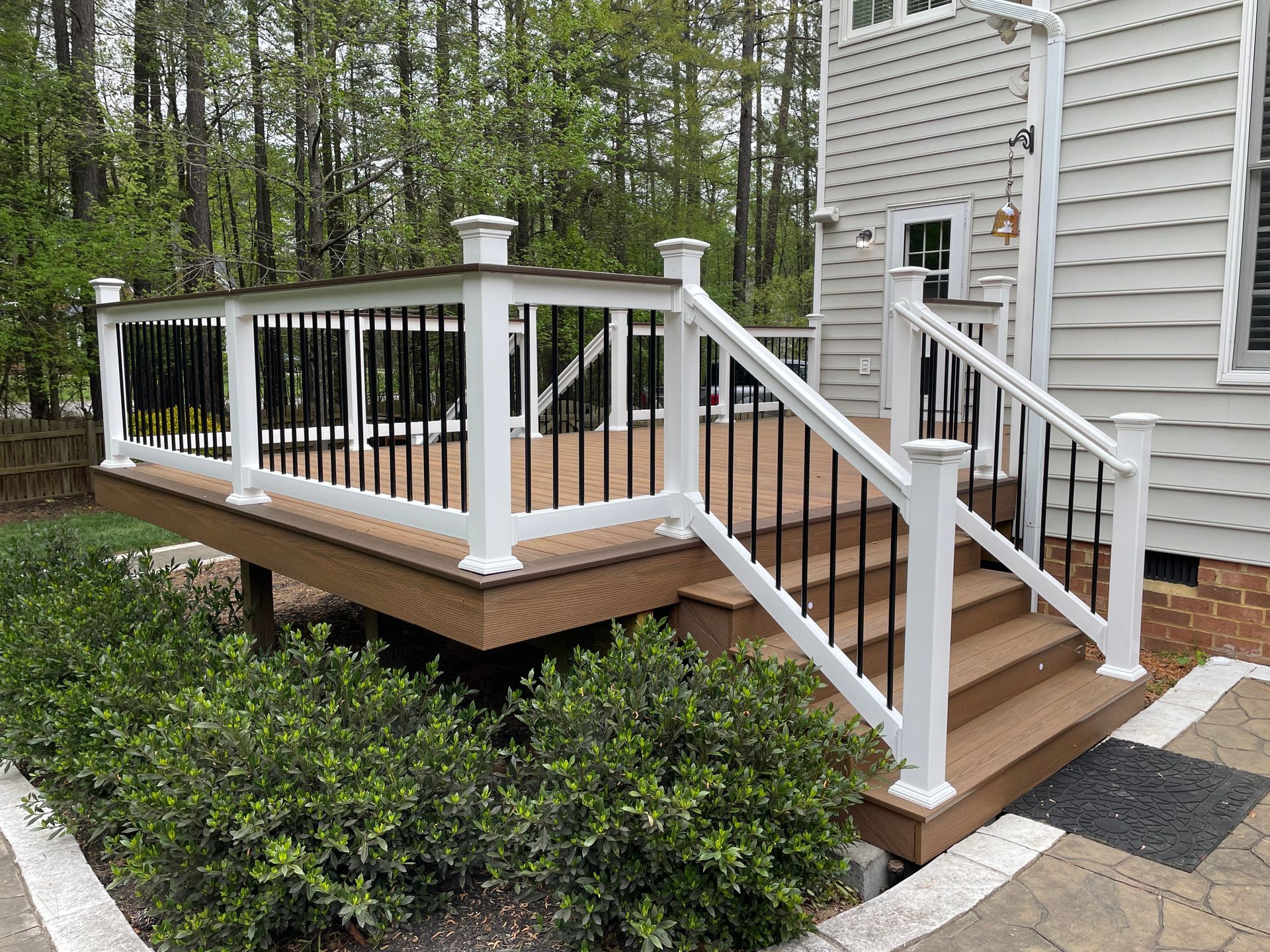 Wooden deck with white railings and steps beside a house, surrounded by trees and shrubs