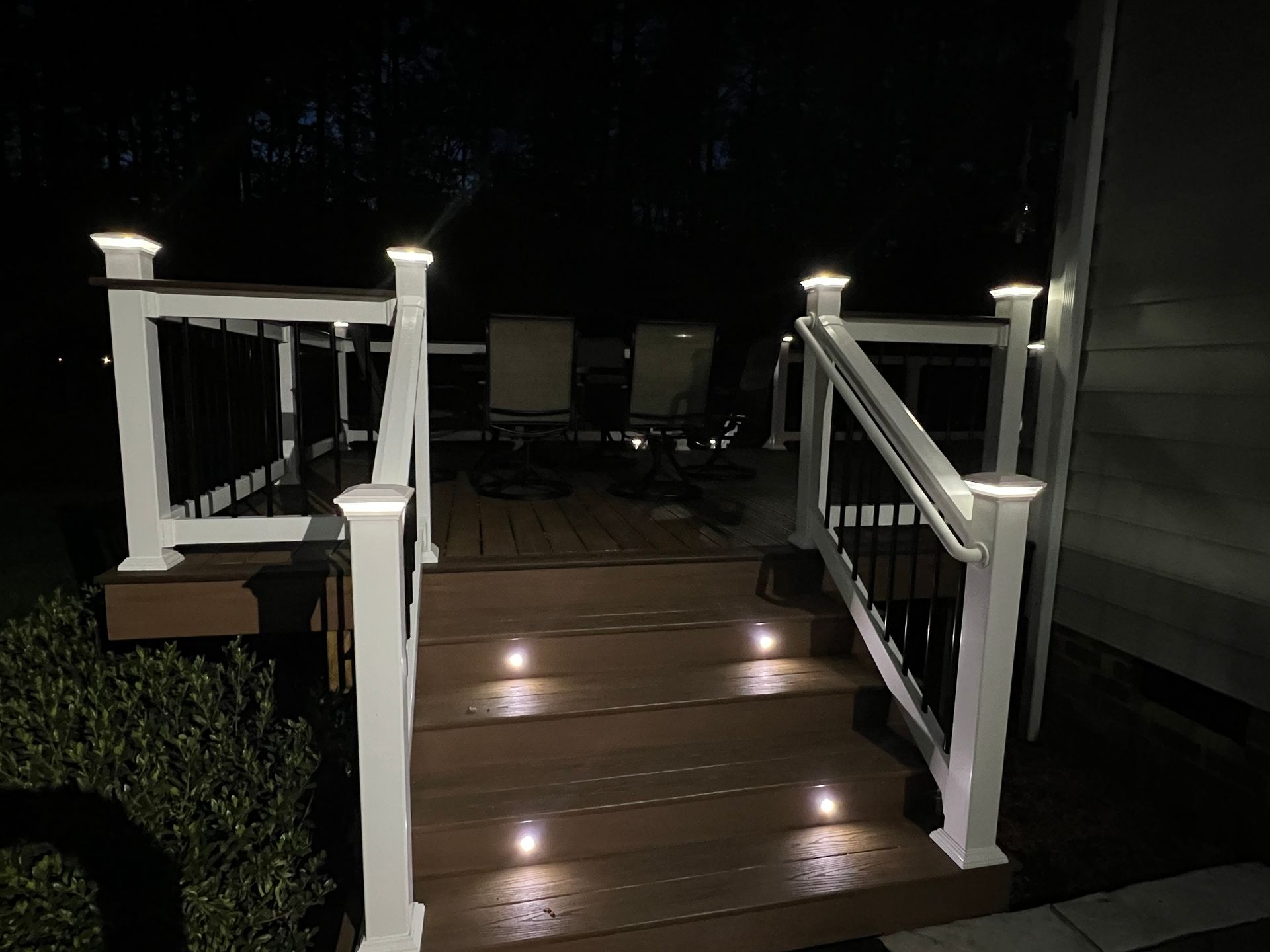 Wooden deck steps with white railings and small lights, leading to outdoor patio chairs at night
